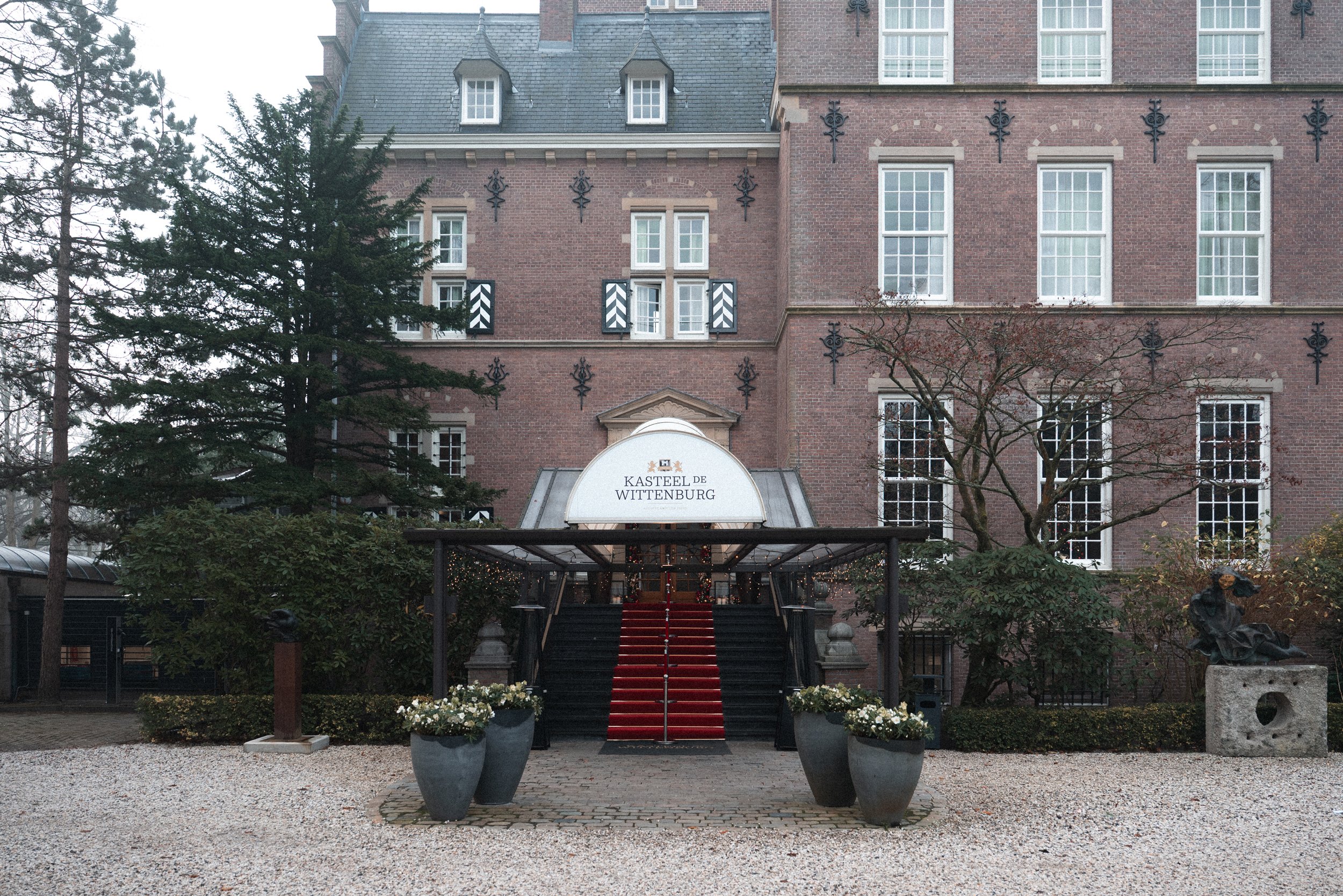 Entrance to Kasteel de Wittenburg, a red brick castle with a staircase leading up to the door, flanked by large planters with flowers and a statue on the right side.