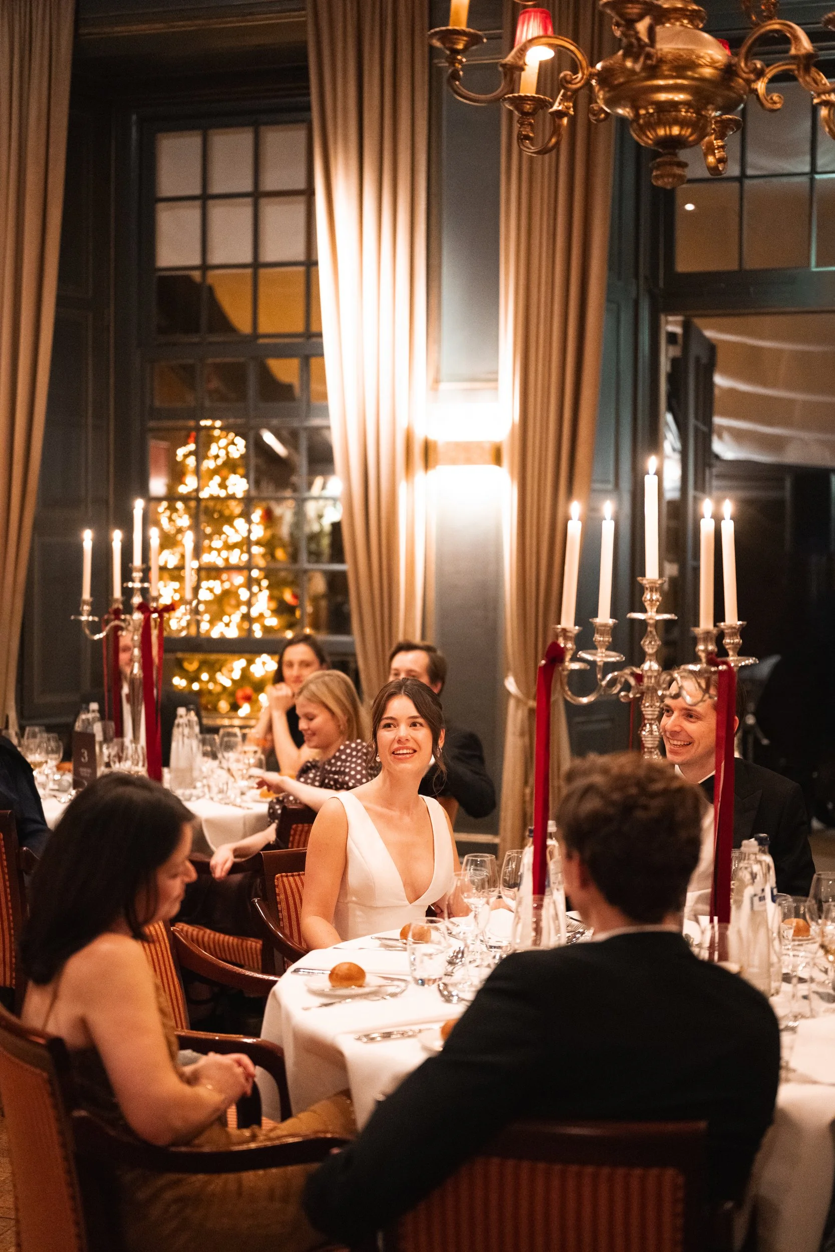 People sitting at a decorated dining table during a festive celebration with a Christmas tree in the background, lit candles, and elegant decor.