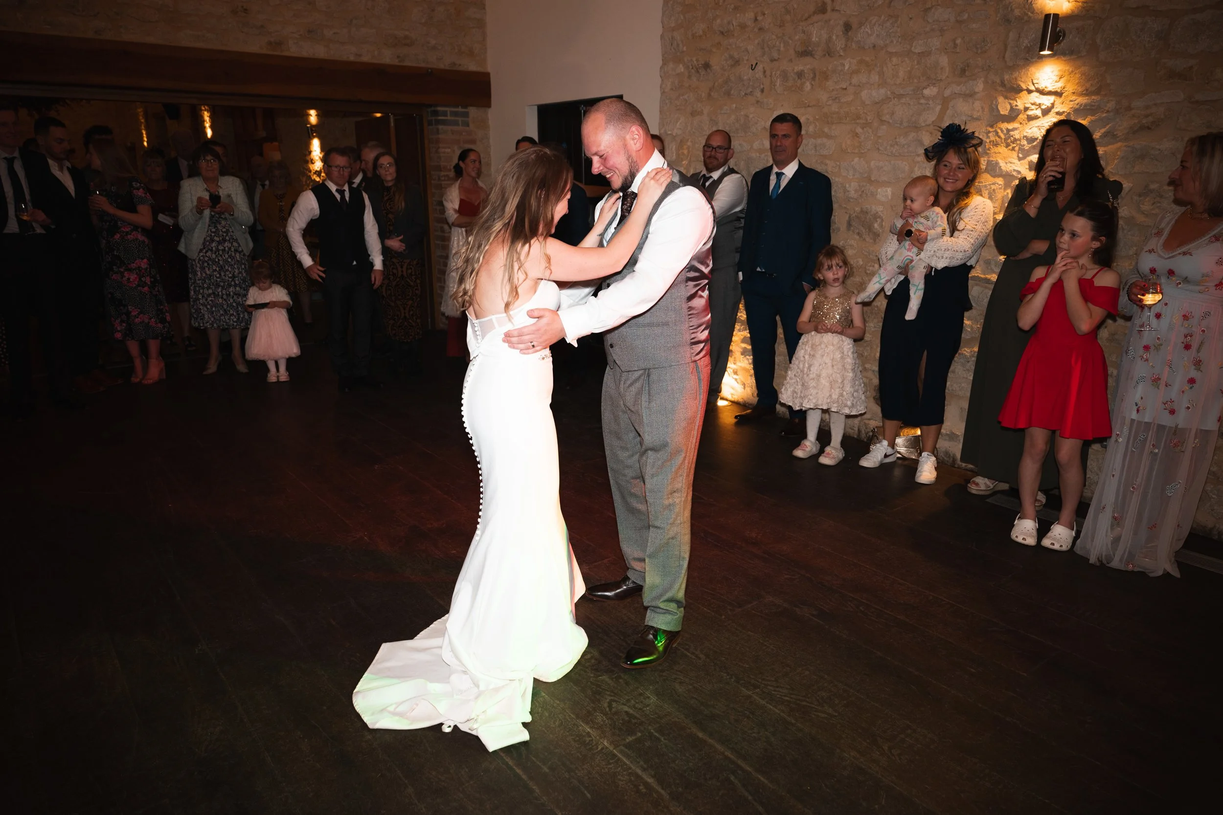 A bride and groom are dancing together at their wedding reception, surrounded by guests who watch and take photos.