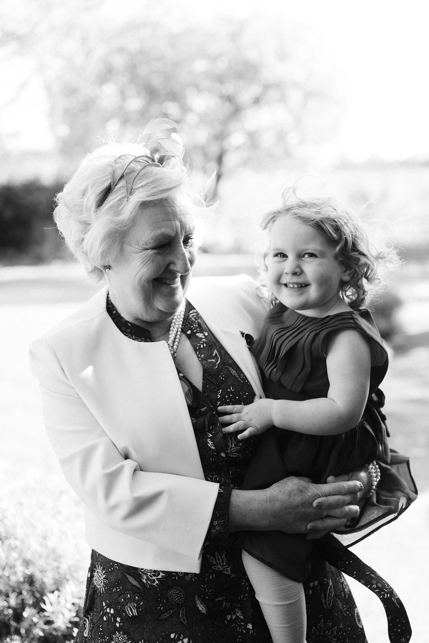 An elderly woman holding a young girl in an outdoor park setting, both smiling and enjoying each other's company.