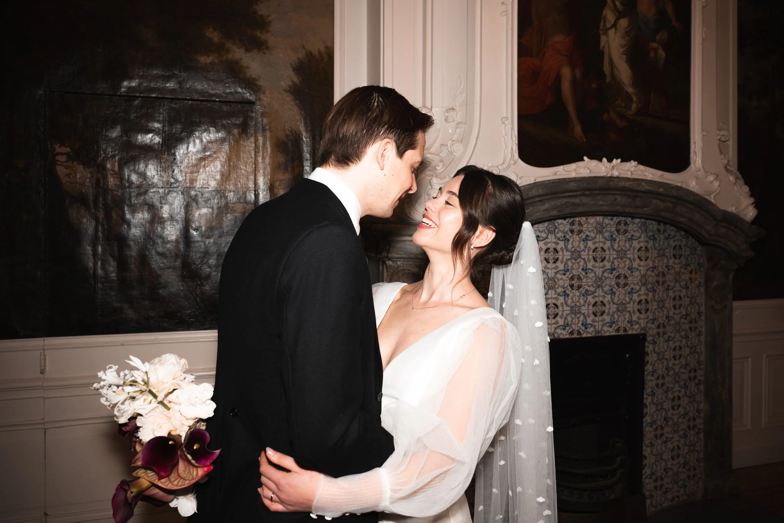 A bride and groom sharing a joyful moment indoors, with the bride holding a bouquet of white and purple flowers, and both smiling at each other.