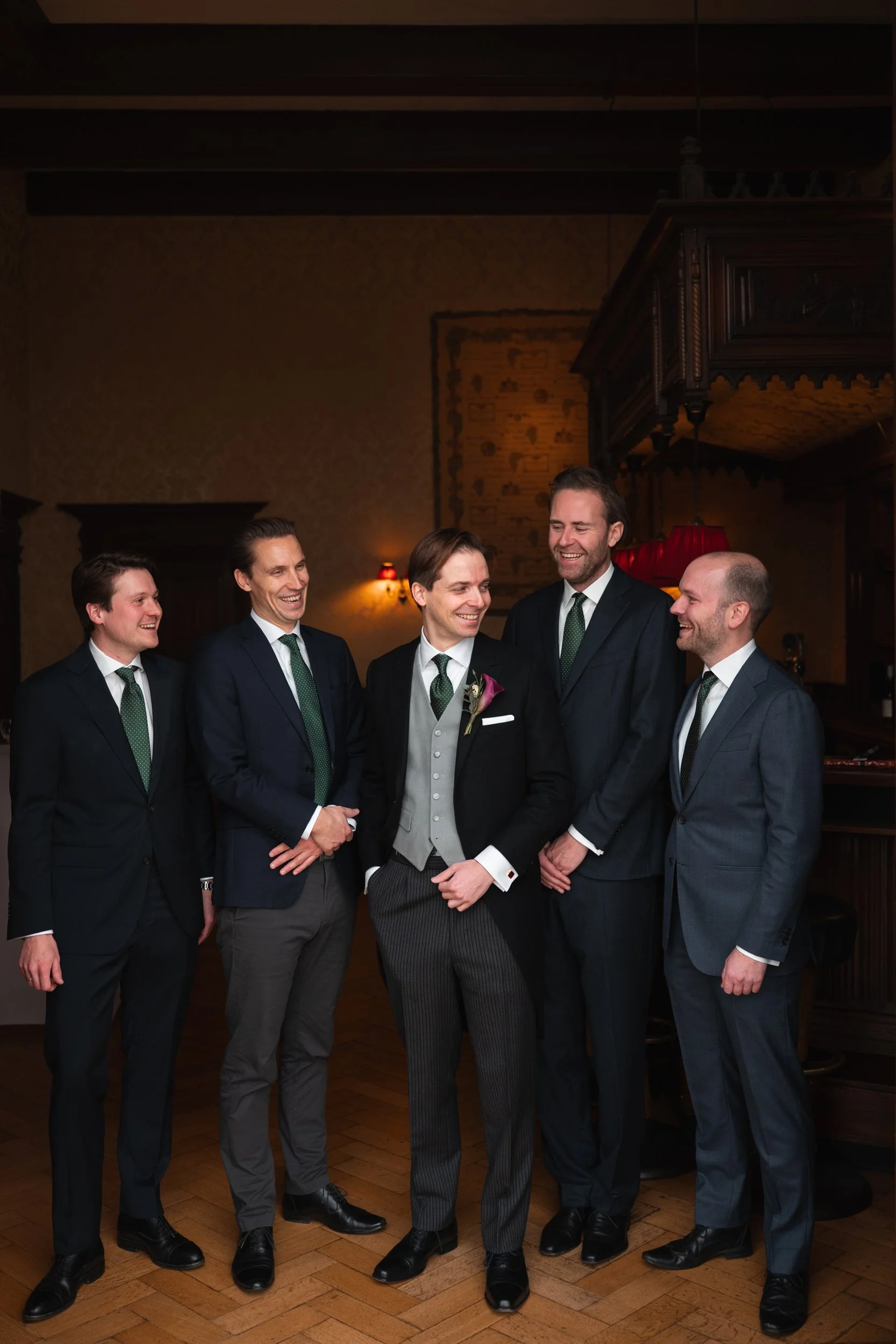 Six men dressed in suits standing together at a social gathering, smiling, in a warmly lit indoor setting with wooden decor, wallpaper, and chandelier.