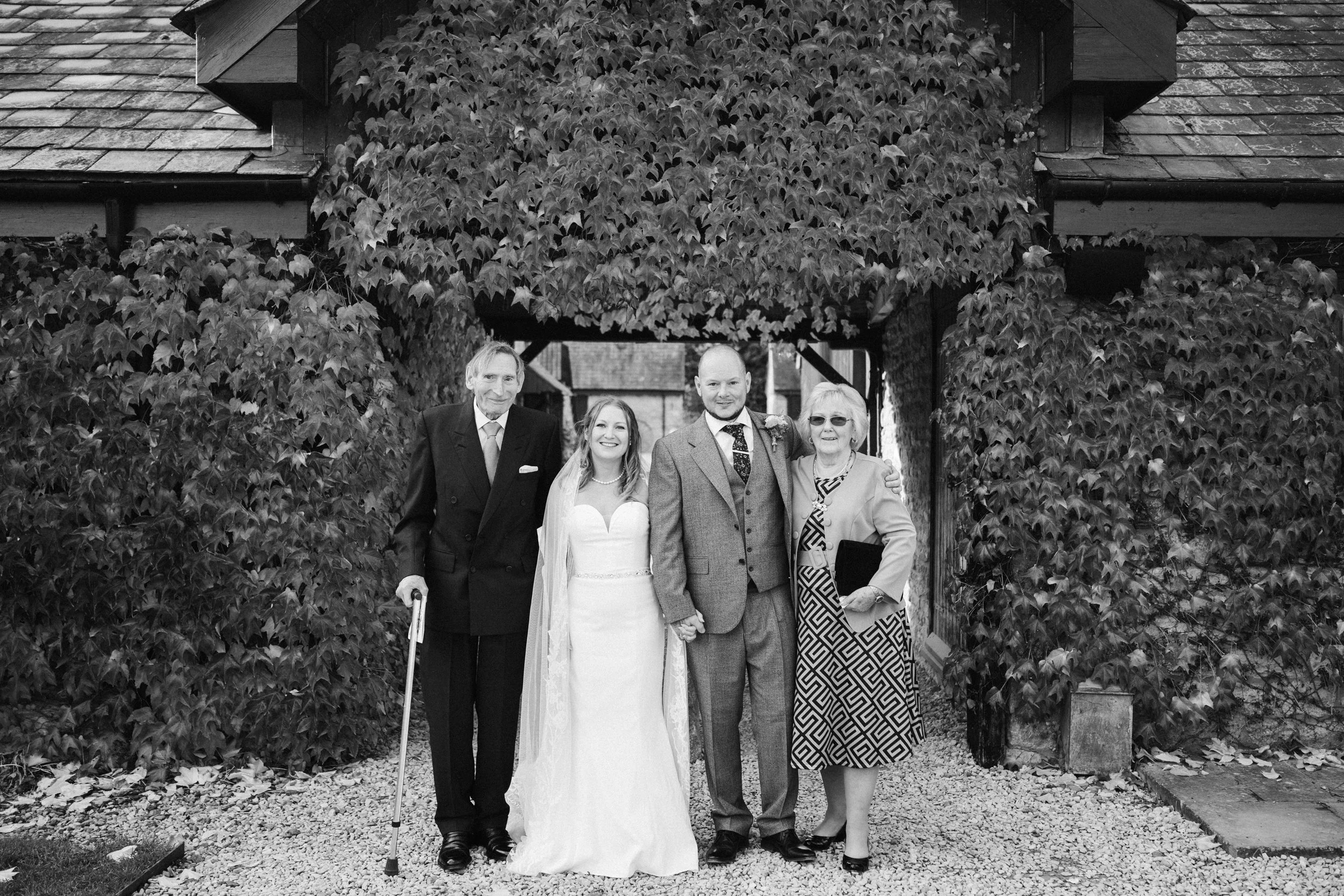 A black-and-white photo of four people standing together outdoors, with one person in a wedding dress, on a gravel path in front of a house with ivy-covered walls.