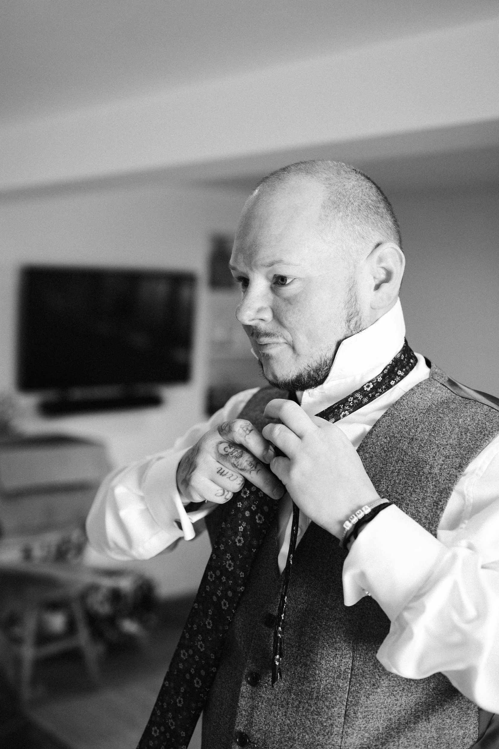A man with tattoos on his fingers, dressed in a vest and shirt, is adjusting his tie in a room with a television in the background.