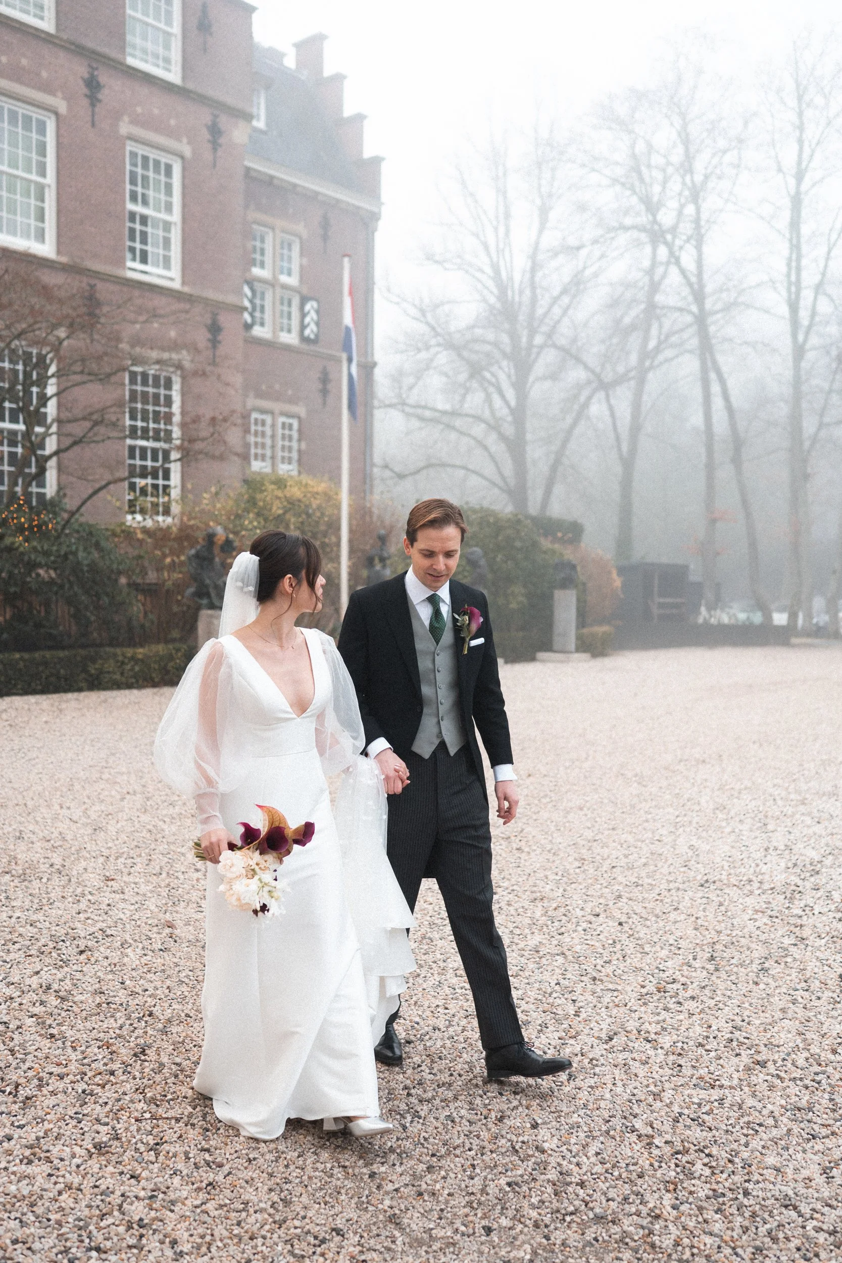 A bride and groom walking hand in hand outside on a foggy day. The bride is wearing a white wedding gown with puffed sleeves and holding a bouquet. The groom is dressed in a formal black suit with a vest and tie. They are walking on a gravel path nea