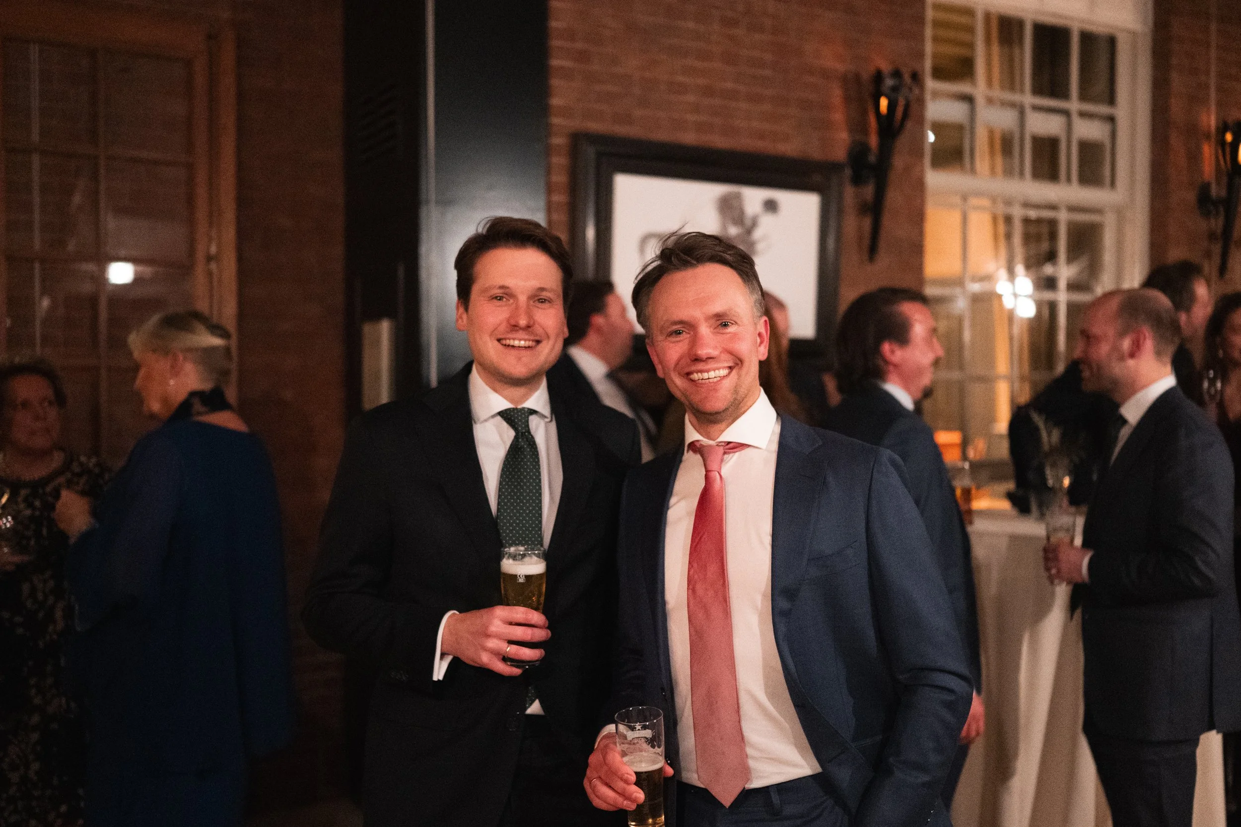 Two men in suits smiling and holding glasses of beer at a social event.