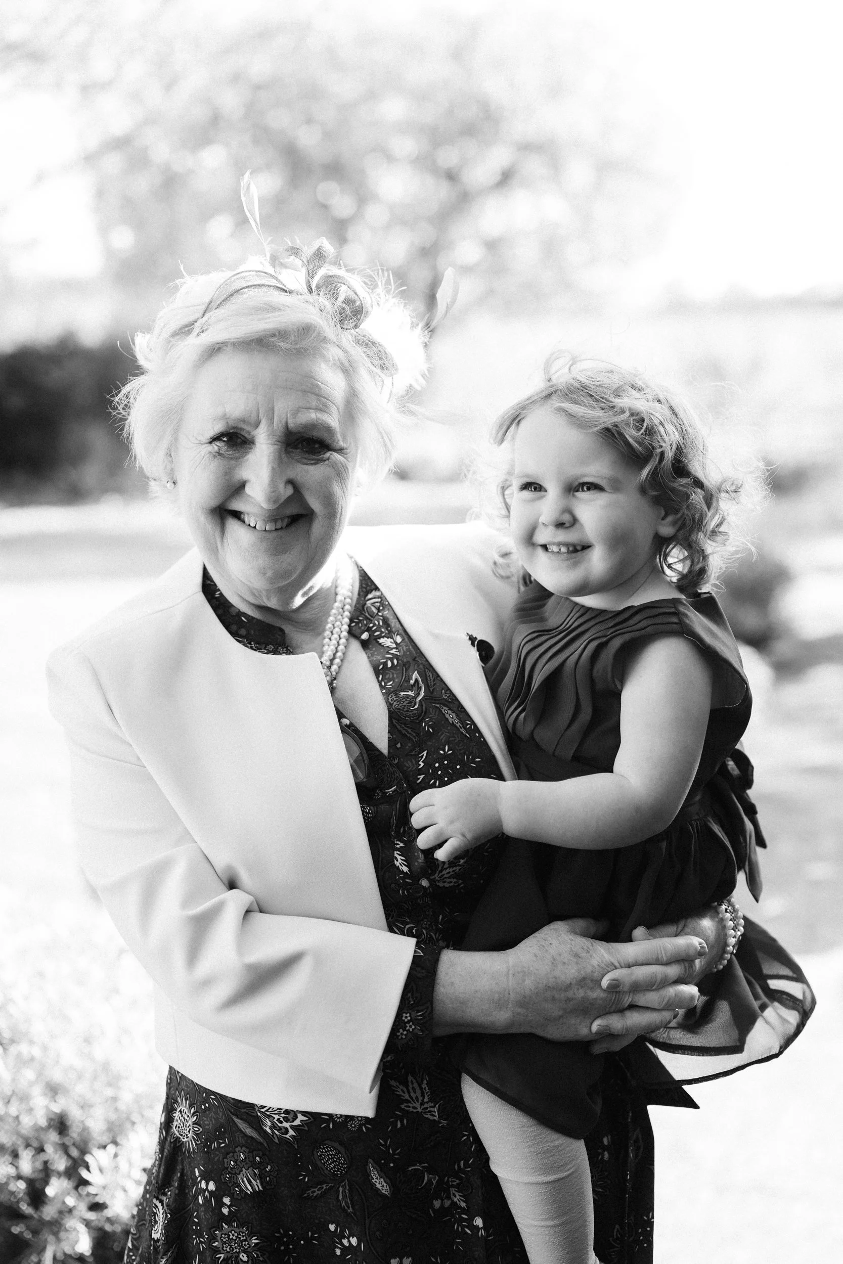 A black and white photo of an elderly woman holding a young girl outdoors, both smiling, with trees and a bright sky in the background.