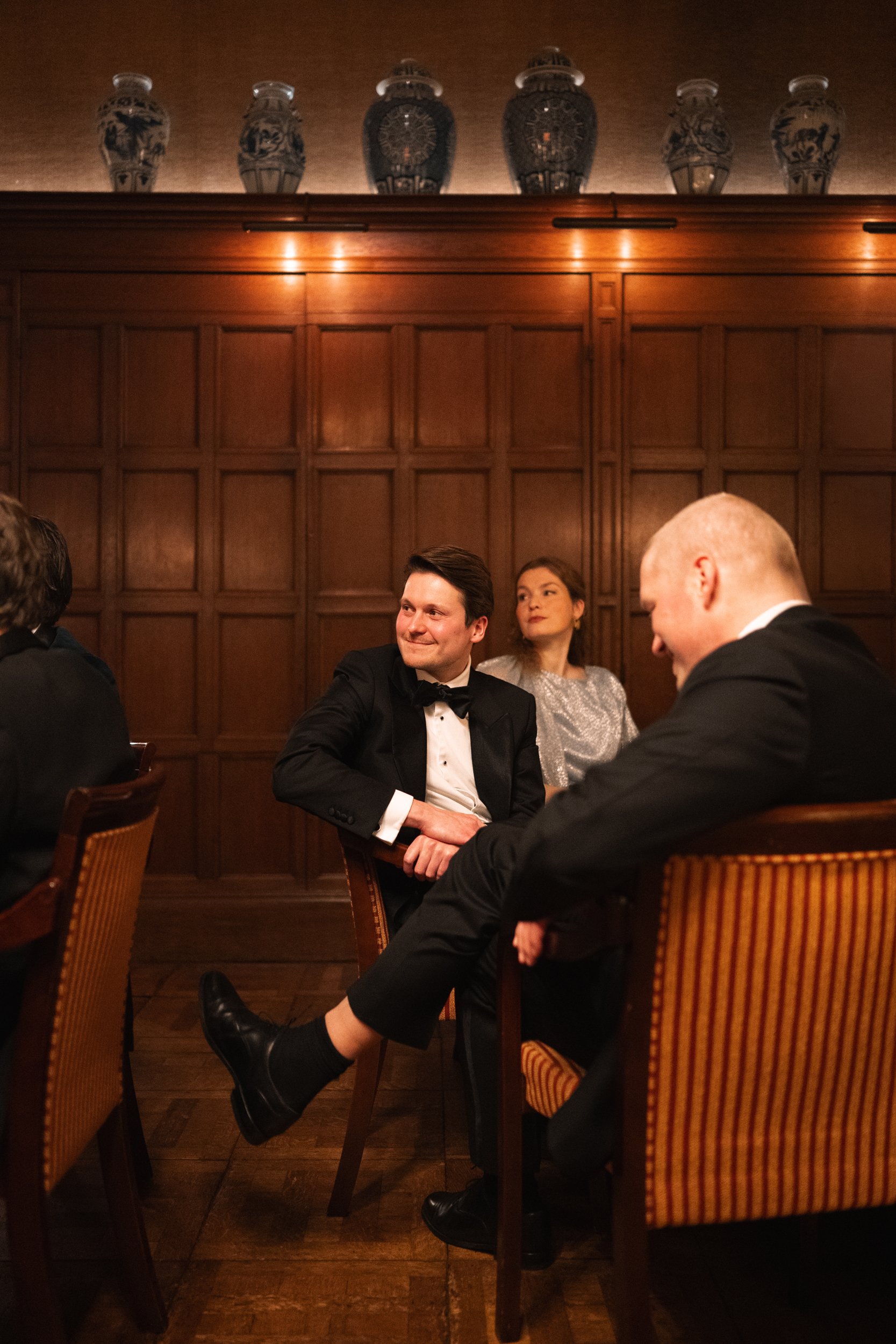People dressed in formal attire dining in an elegant room with wooden paneling and decorative vases on the shelf above.