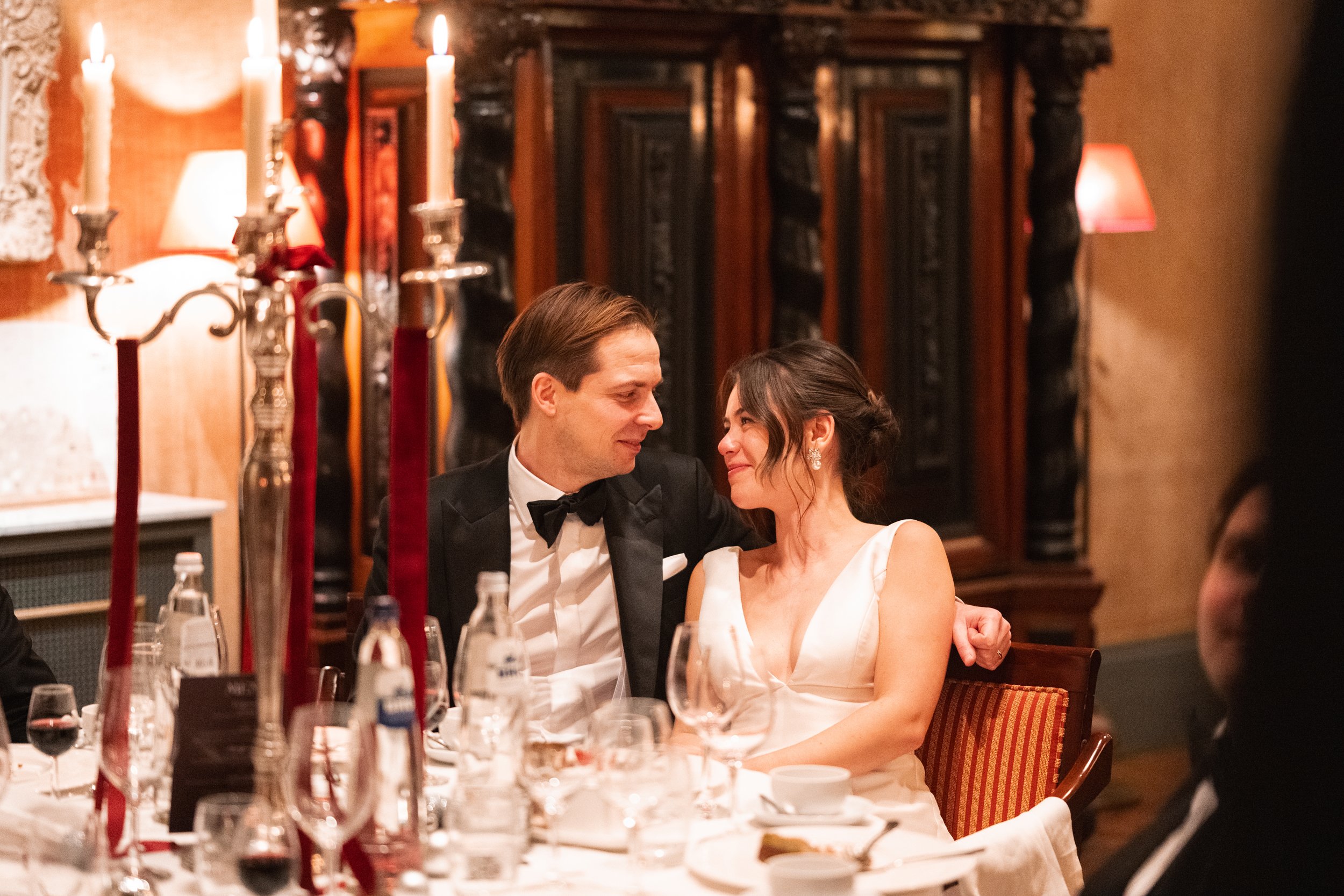 A man and woman dressed formally are sitting close together at a dinner table, gazing into each other's eyes in a warmly lit, elegant restaurant or banquet hall.
