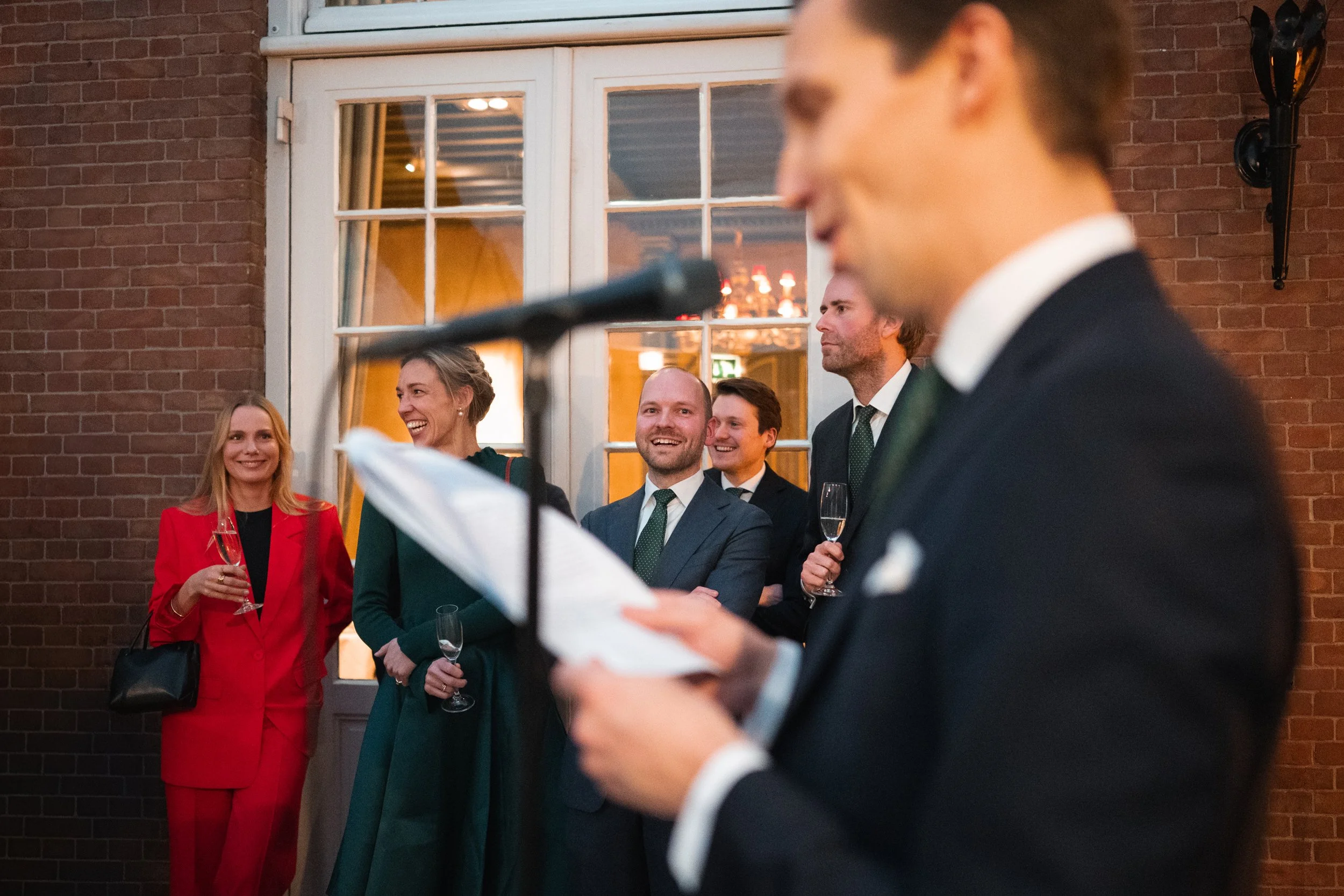 A man in a dark suit is reading from a paper at a microphone, with a group of smiling people in formal attire, holding champagne glasses, listening in the background.