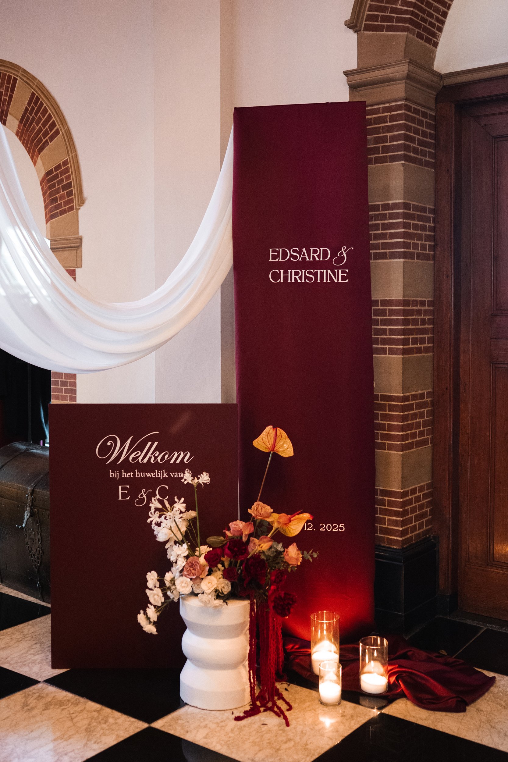 Wedding decoration with burgundy and white signs, floral arrangement in a white vase, and lit candles on a checkered floor.