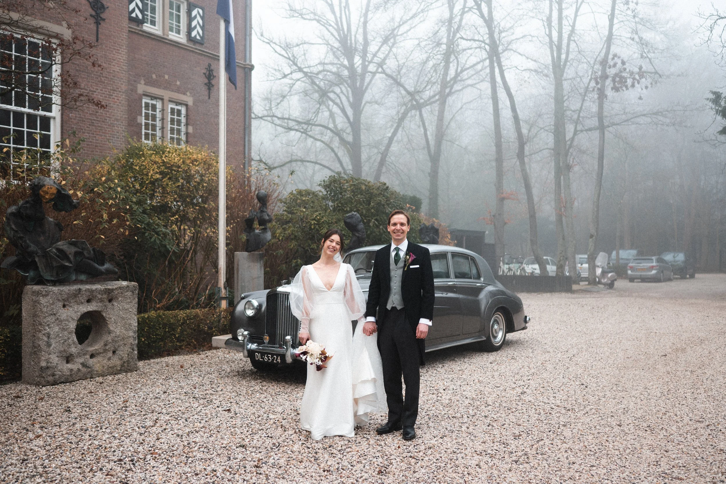 A newlywed couple stands on a gravel driveway, holding hands and smiling. The bride wears a white wedding dress and holds a bouquet, while the groom wears a black tuxedo with a gray vest. Behind them is a vintage black car, and the background features a building with art sculptures, bushes, and leafless trees shrouded in fog.