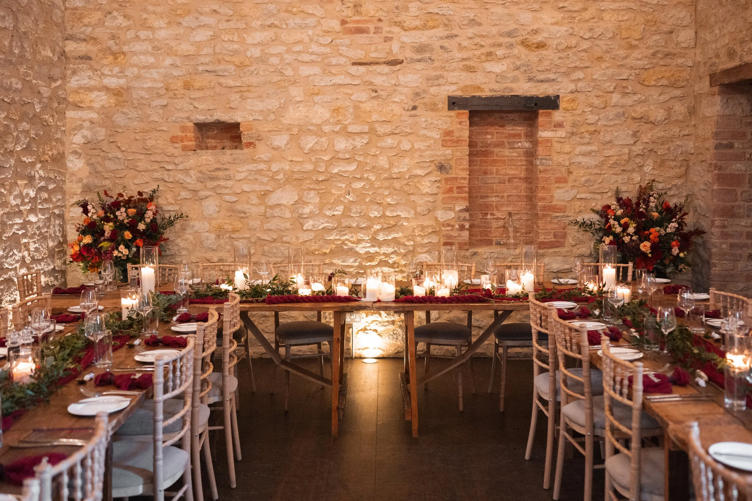 Elegant dining table decorated with candles, greenery, and red accents, set for a celebration in a rustic stone-walled room with floral arrangements at both ends.
