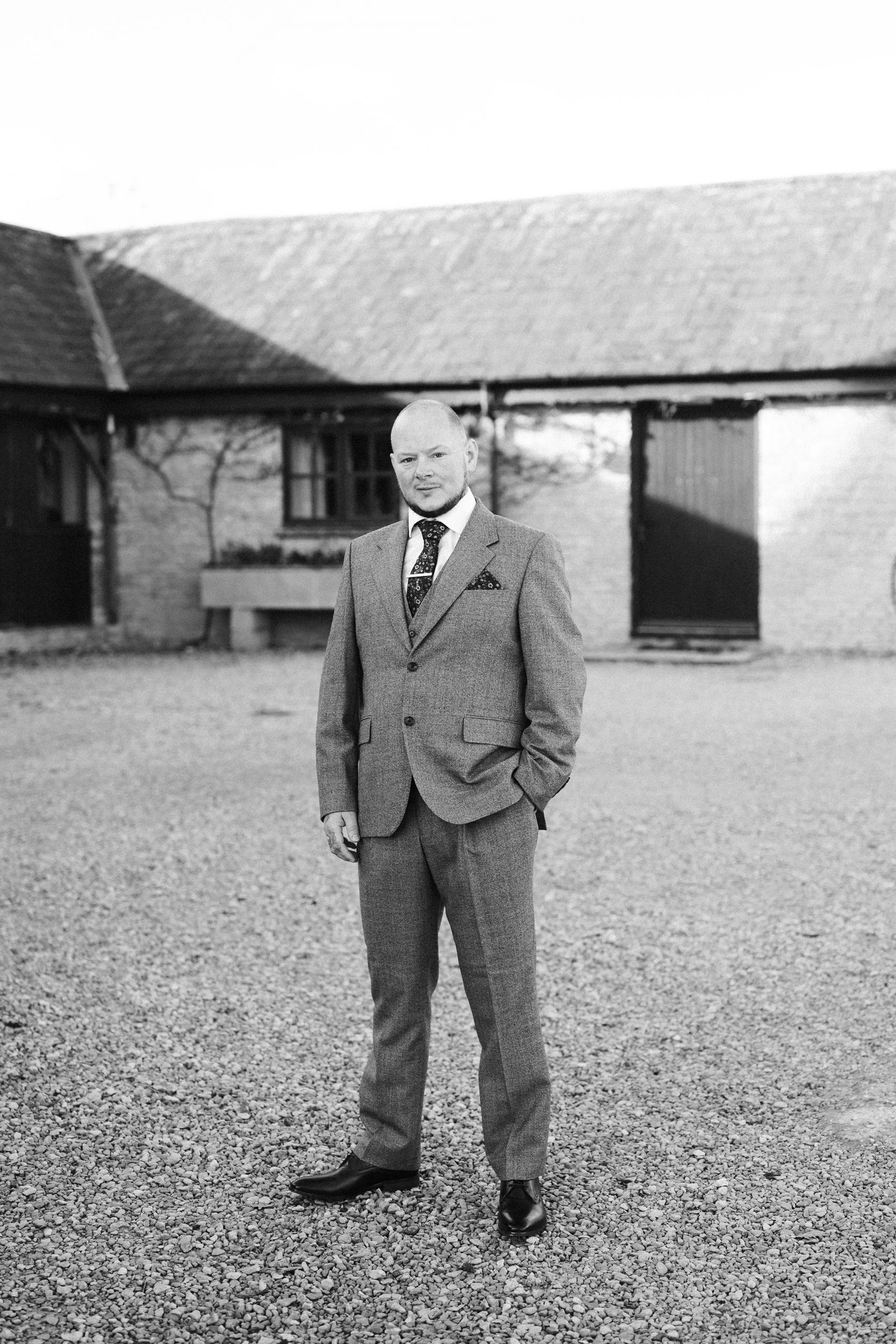 A man in a suit standing outside on gravel with a building in the background.