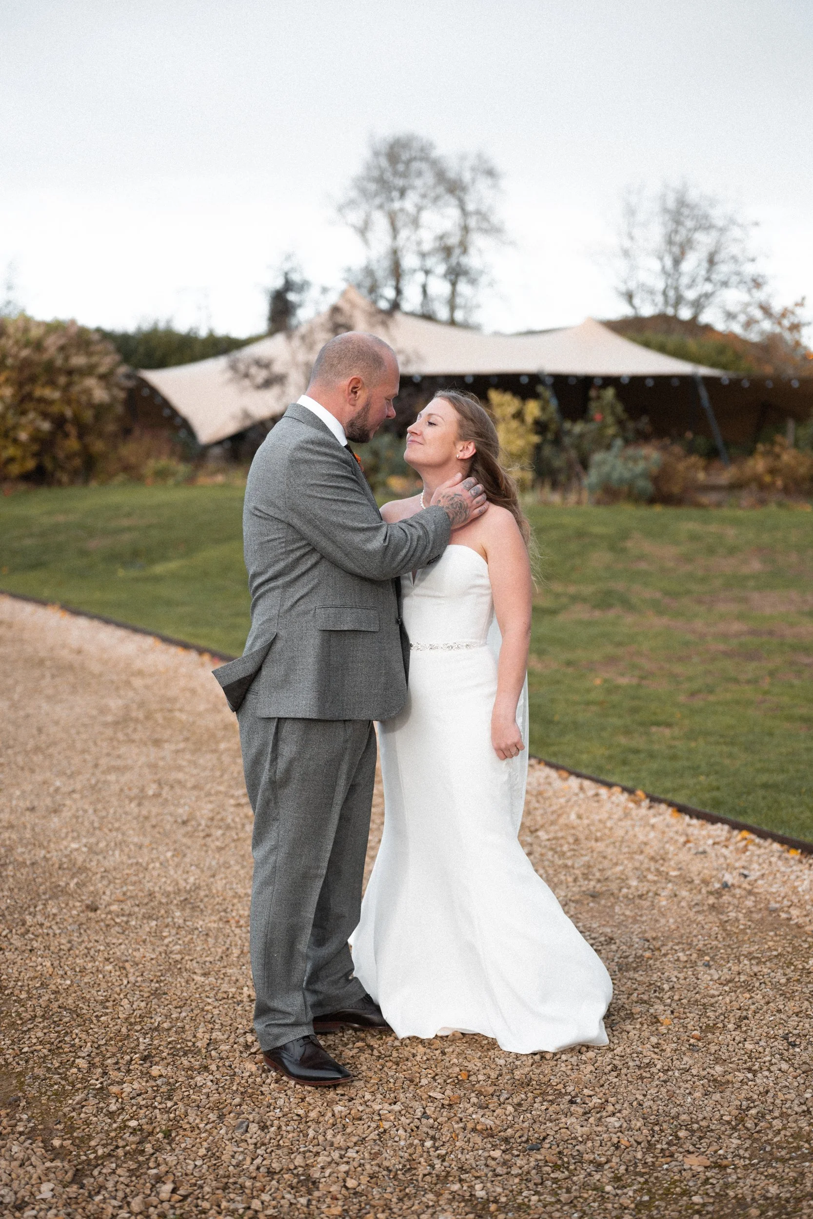A bride and groom sharing an emotional moment during their wedding outdoors, with the bride in a white dress and the groom in a gray suit, standing on a gravel path with a landscaped garden and tents in the background.