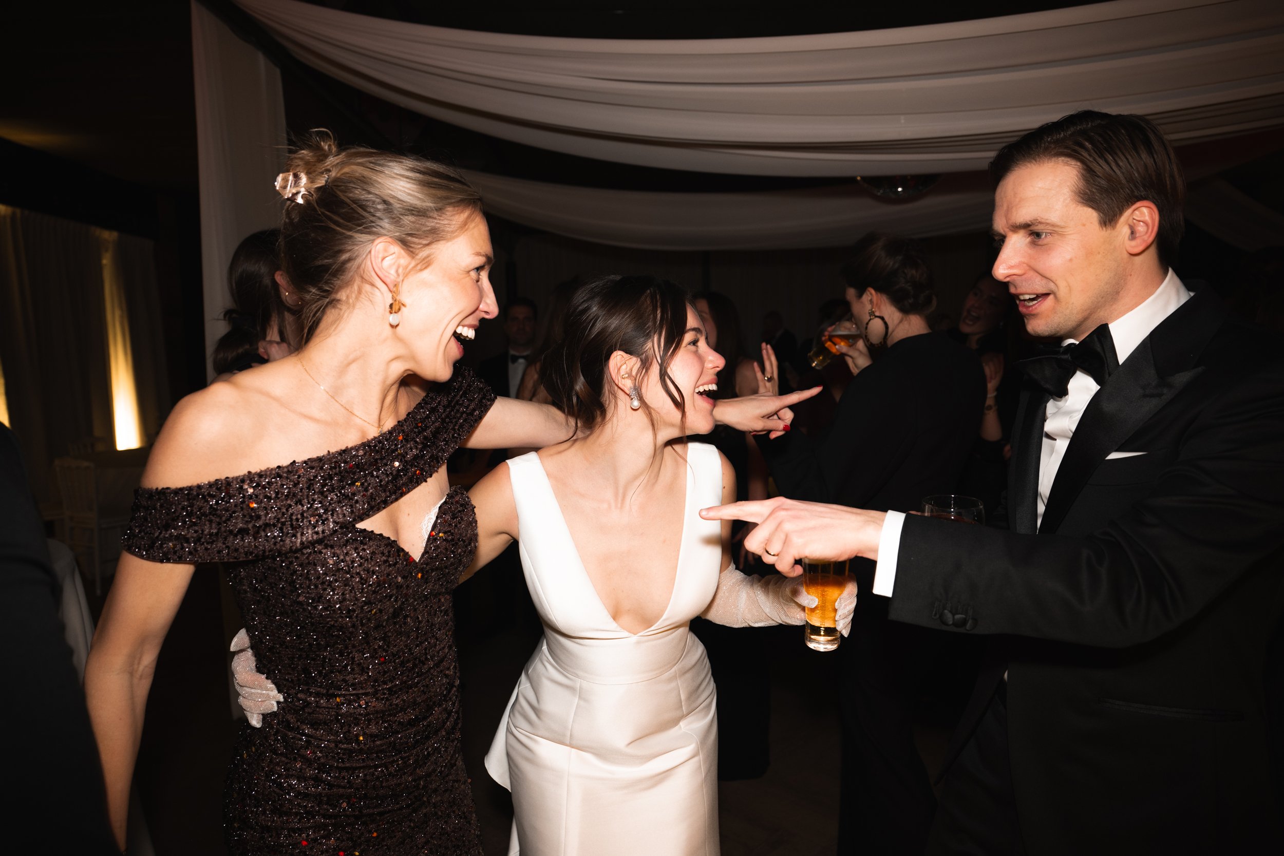 People at a formal celebration or wedding dancing and laughing, including a woman in a dark sequined dress, a woman in a white dress, and a man in a tuxedo.