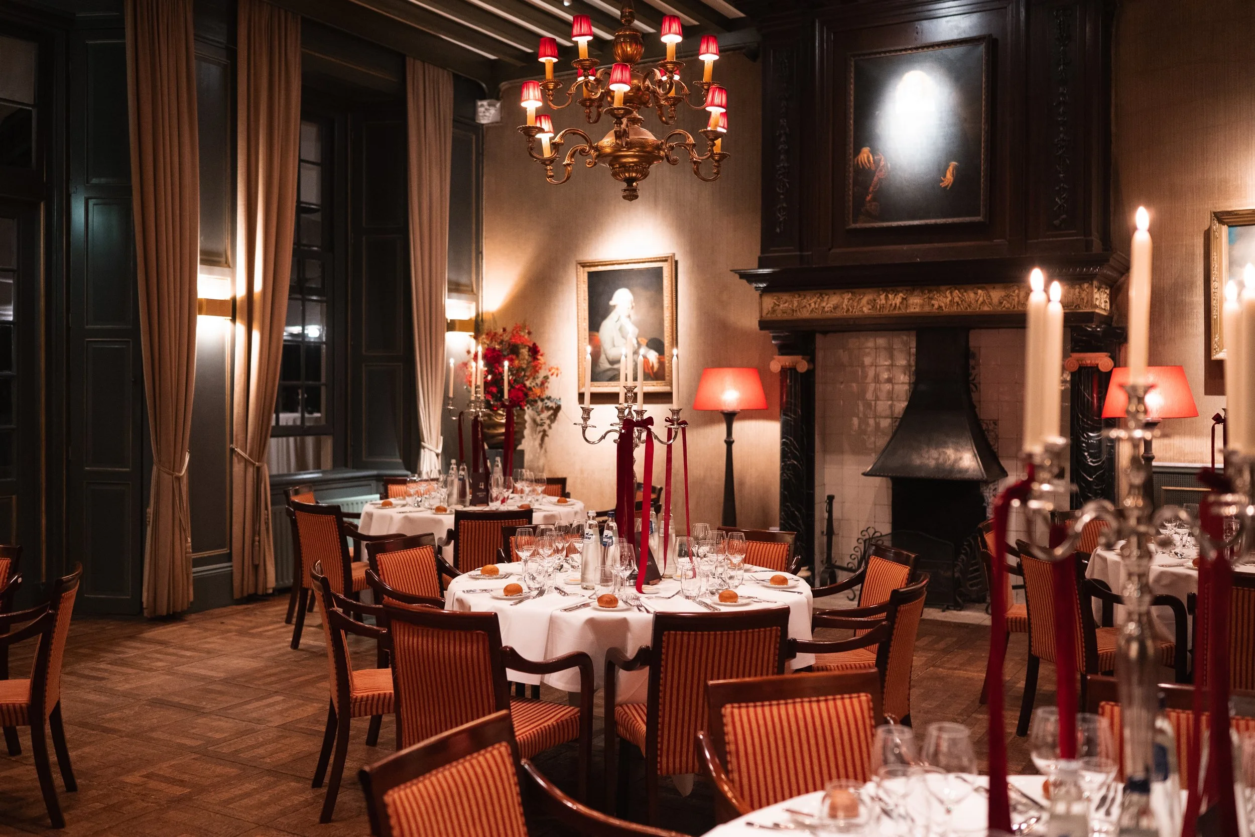An elegant dining room decorated for a special event, featuring round tables with white tablecloths, red and gold striped chairs, tall candles on candelabras, and a chandelier with red lampshades. The room has dark wood paneling, a large fireplace, f