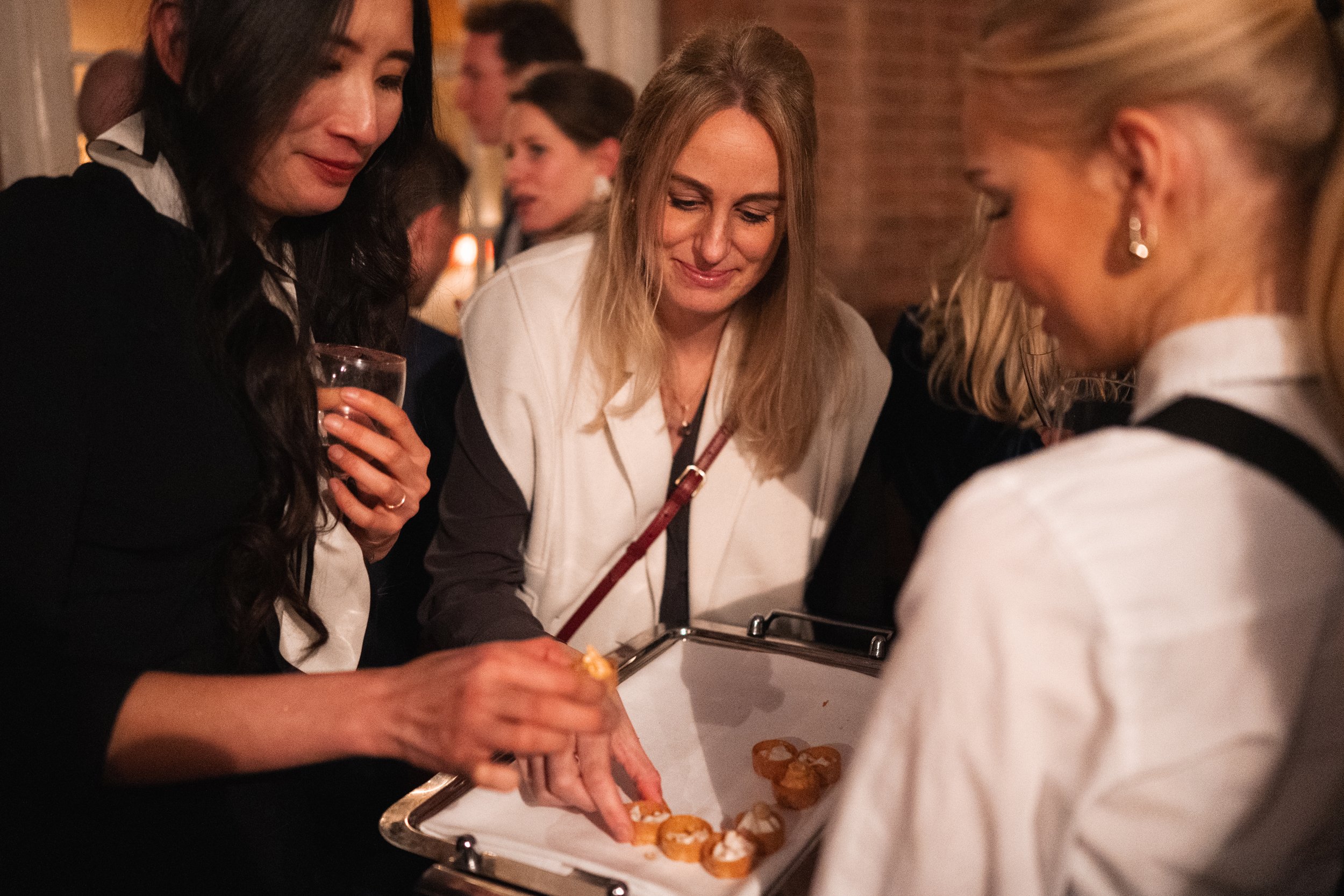 Three women gather around a tray of small desserts or pastries at a social event, with other guests visible in the background.