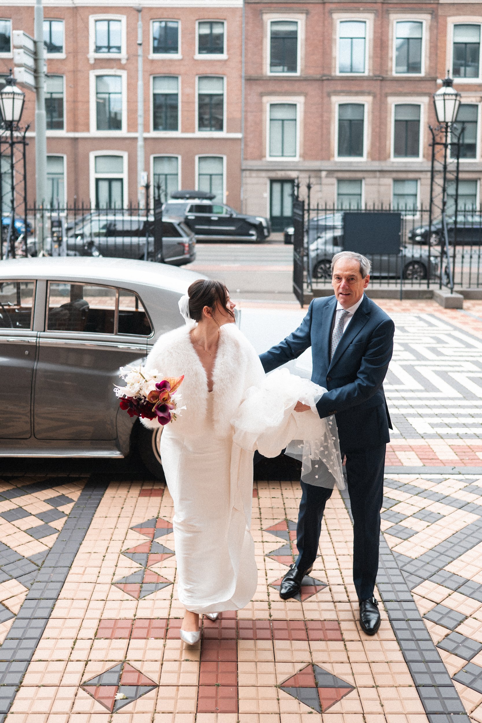 A bride carrying a bouquet and a groom in a suit are exiting a vintage car, with the groom holding the bride's train, outside a building in an urban area.