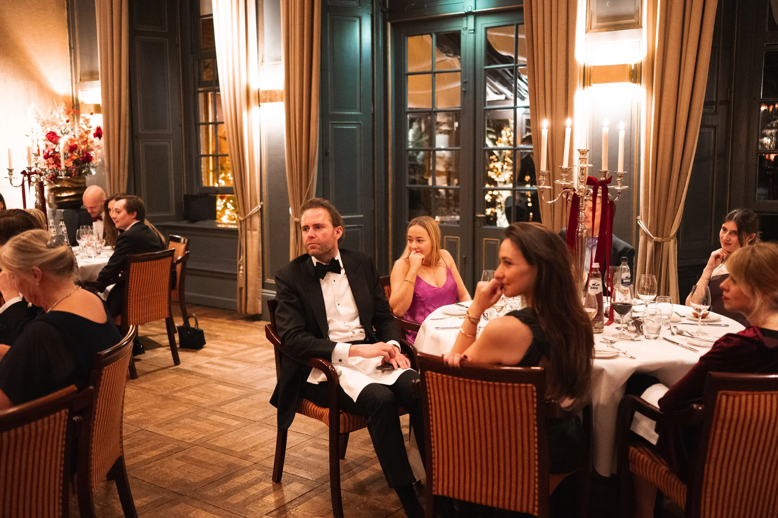 People dressed in formal attire, sitting at tables in a warmly lit dining room decorated for the holidays, with a Christmas tree visible outside the window.