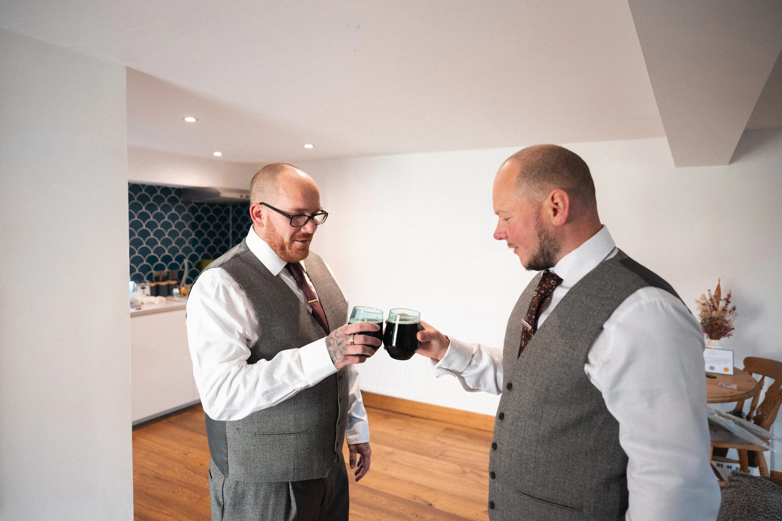 Two men dressed in white shirts and gray vests, with one wearing glasses, clinking glasses filled with dark beverages indoors, with wooden floors and white walls.
