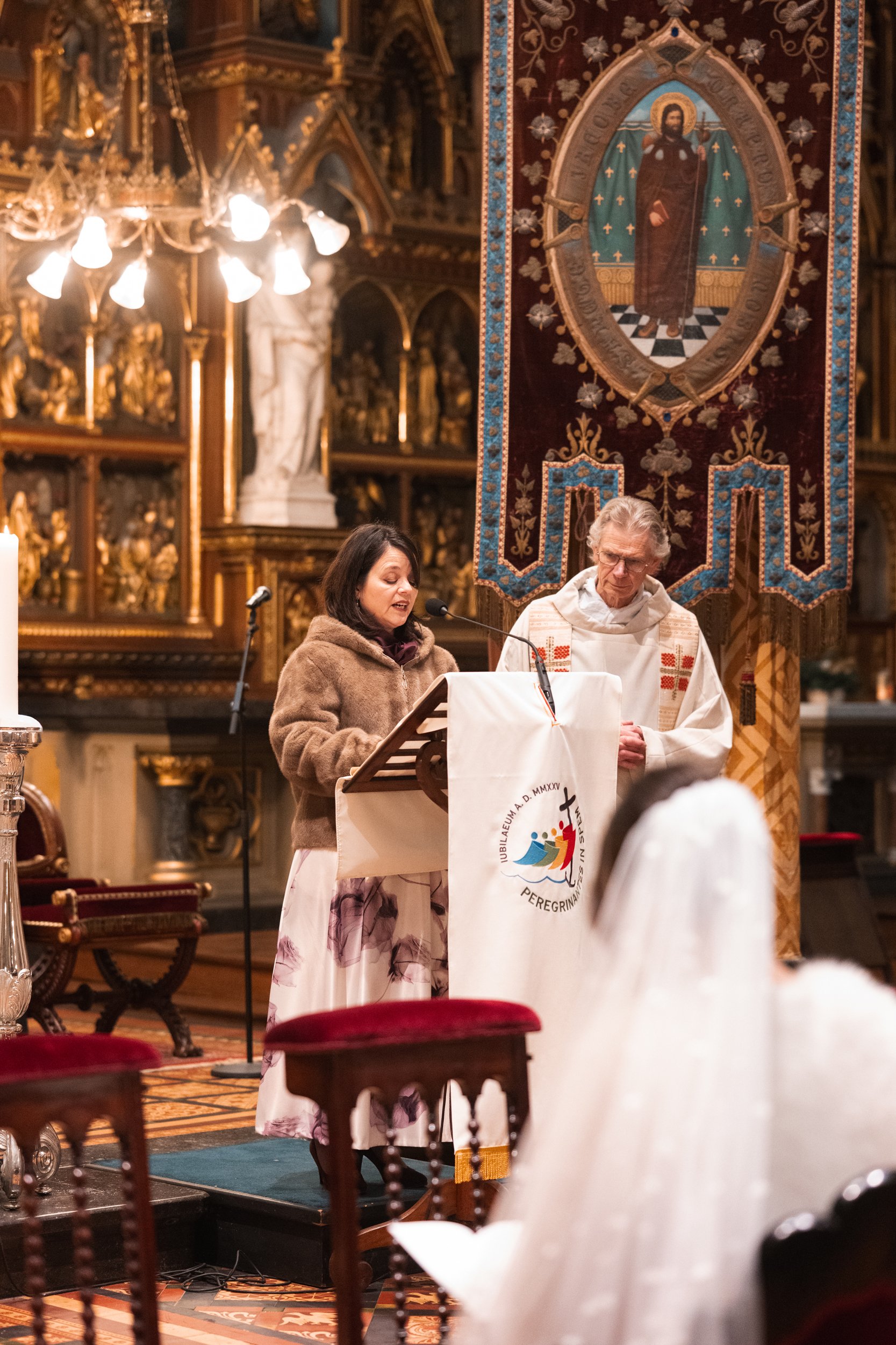 A woman and a priest standing at a lectern during a religious service inside a church, with ornate decorations, statues, and a large banner depicting a saint or religious figure in the background.