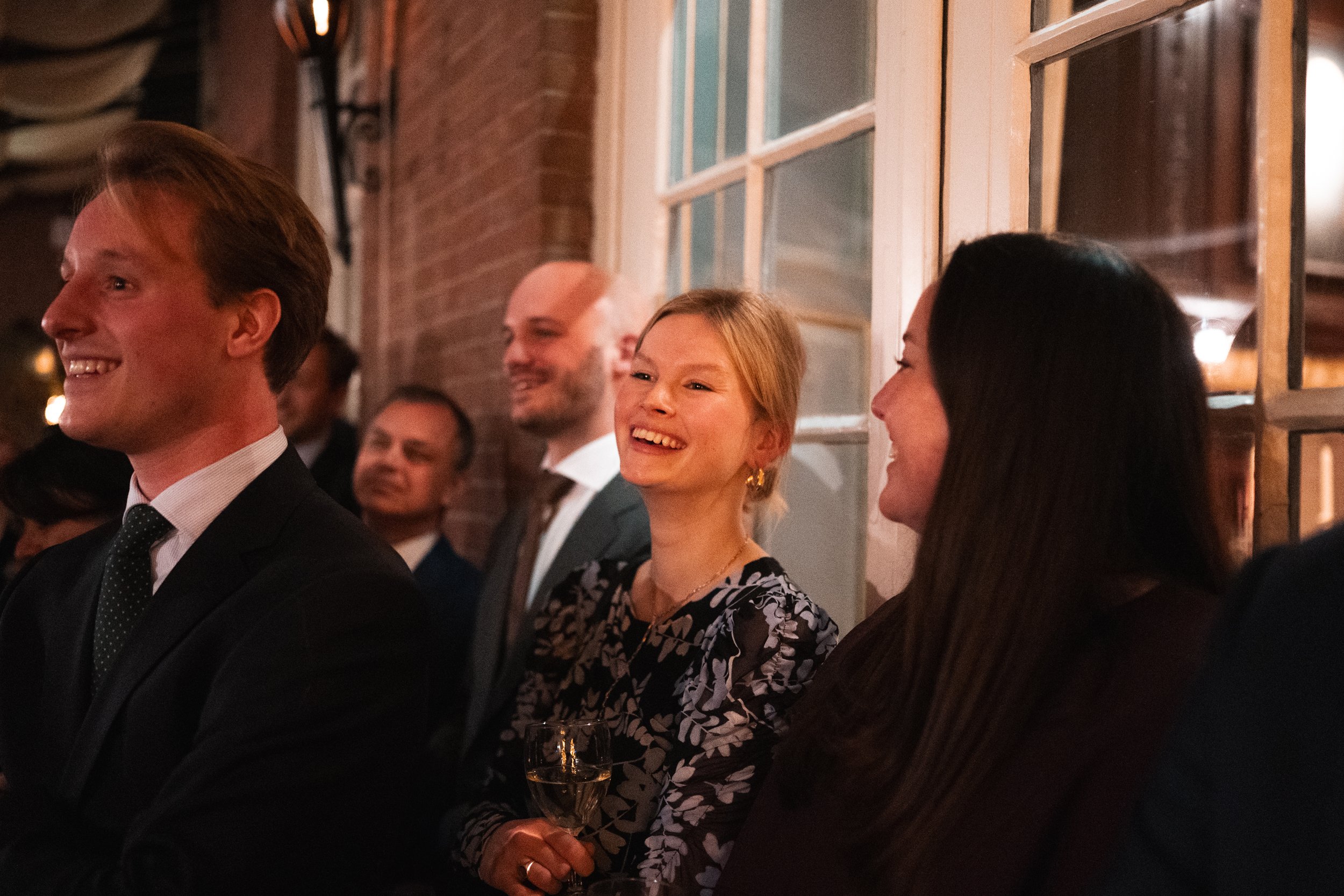 People sitting and enjoying a social event, smiling and laughing, holding glasses of champagne in a warmly lit indoor setting with brick walls and windows.