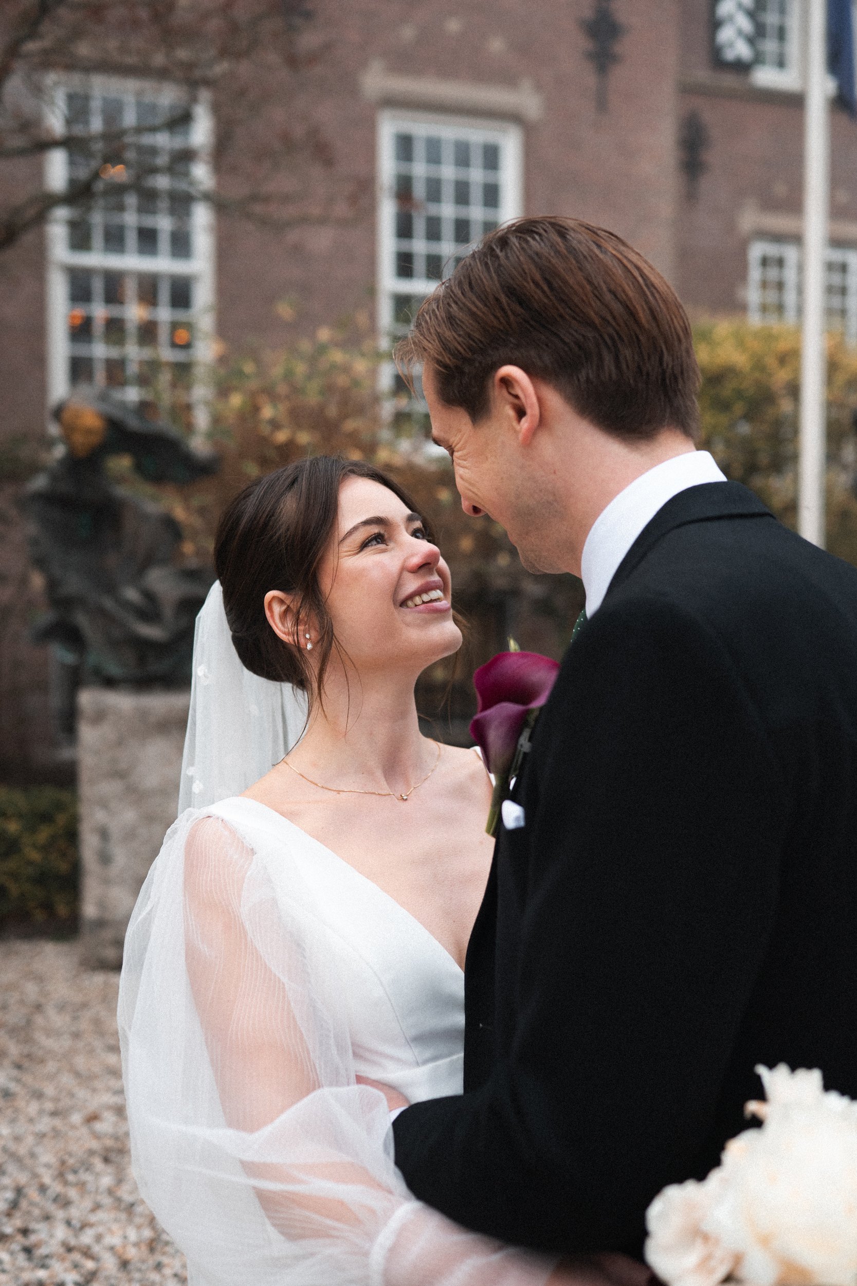 A bride and groom smiling at each other during their wedding outdoors, with a brick building and autumn trees in the background.