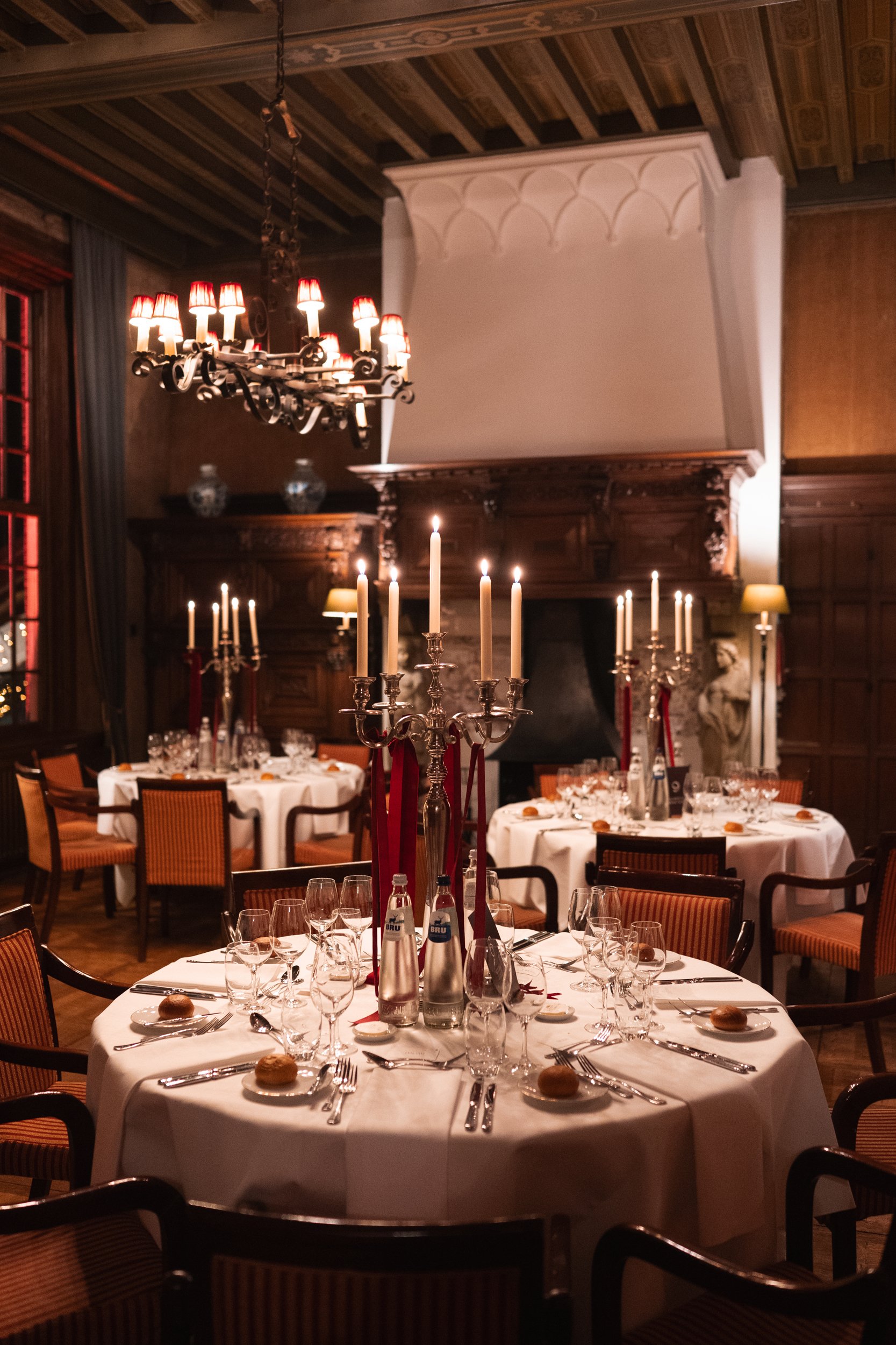 Elegant dining room with round tables set for a formal dinner, featuring candles, glassware, and bread rolls, with a large fireplace, chandeliers, and wooden paneling.