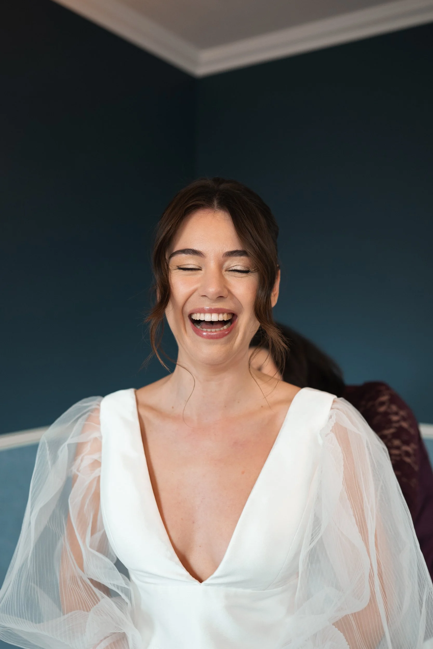 A woman with brown hair laughing, wearing a white dress with sheer sleeves, against a dark blue wall.