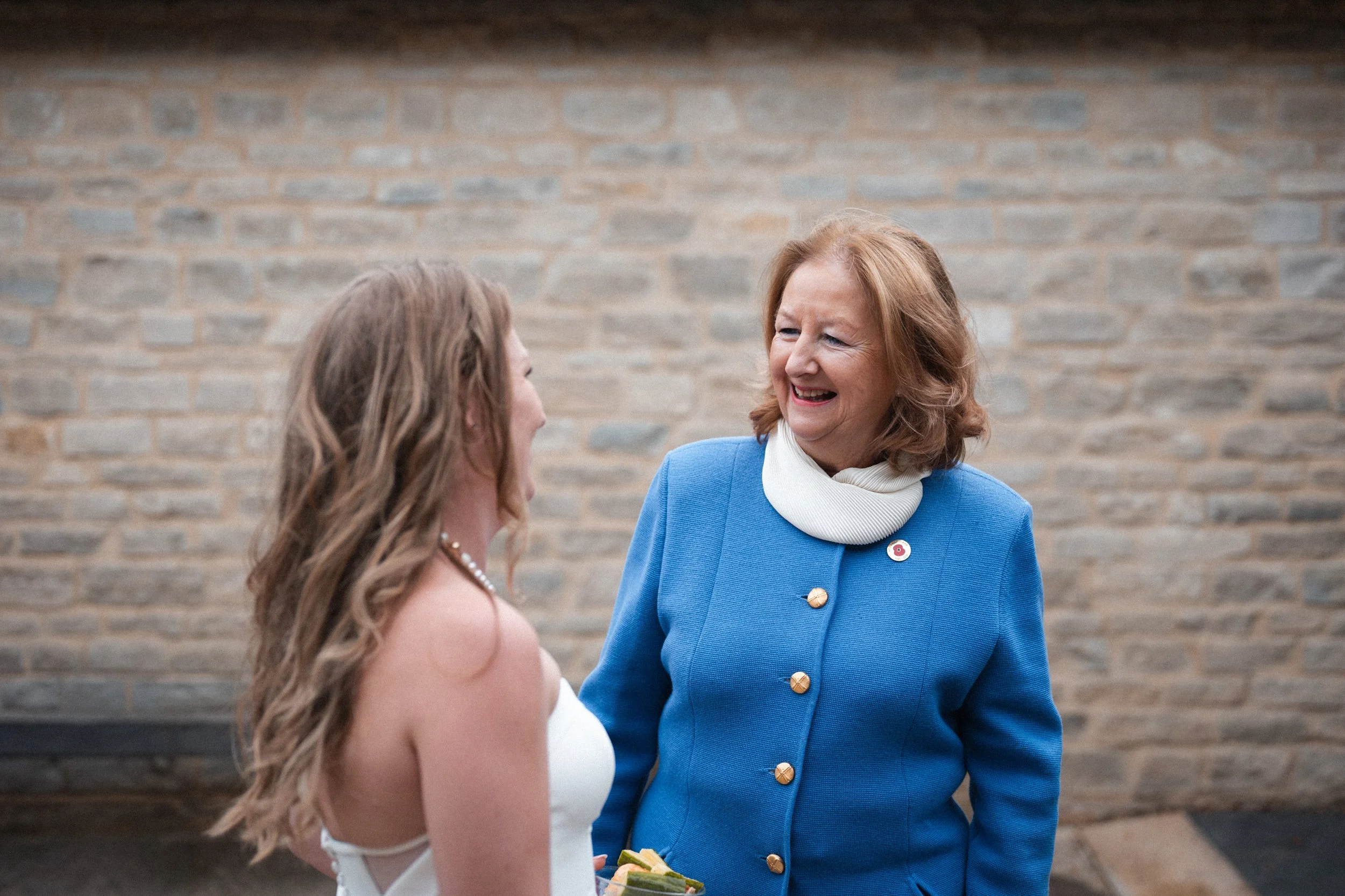 A woman with red hair, wearing a blue jacket with gold buttons and a white turtleneck, smiling and talking to a young woman in a white dress, with a pearl necklace, against a brick wall.