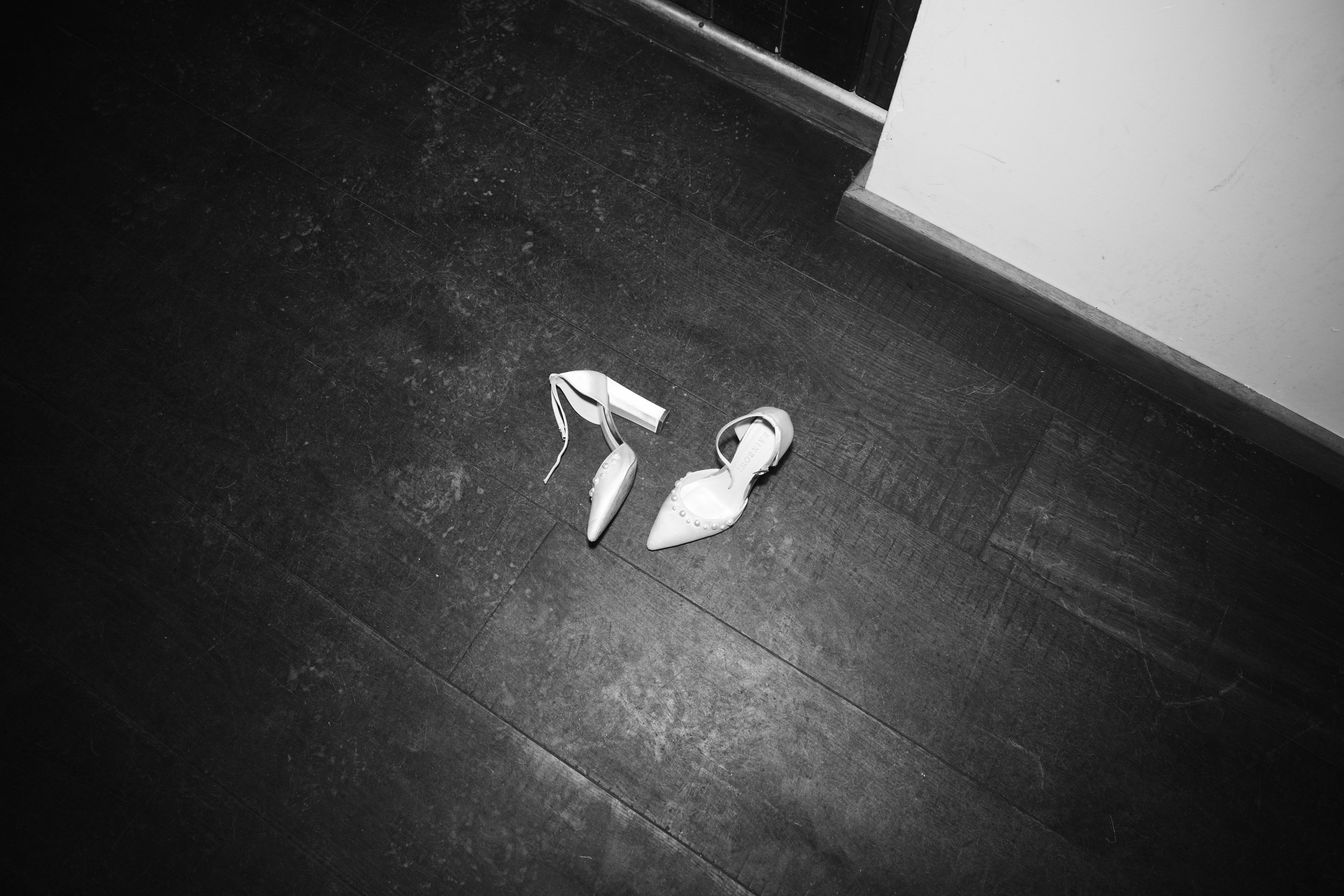 A pair of white high-heeled shoes on a dark wooden floor near a wall and a door.