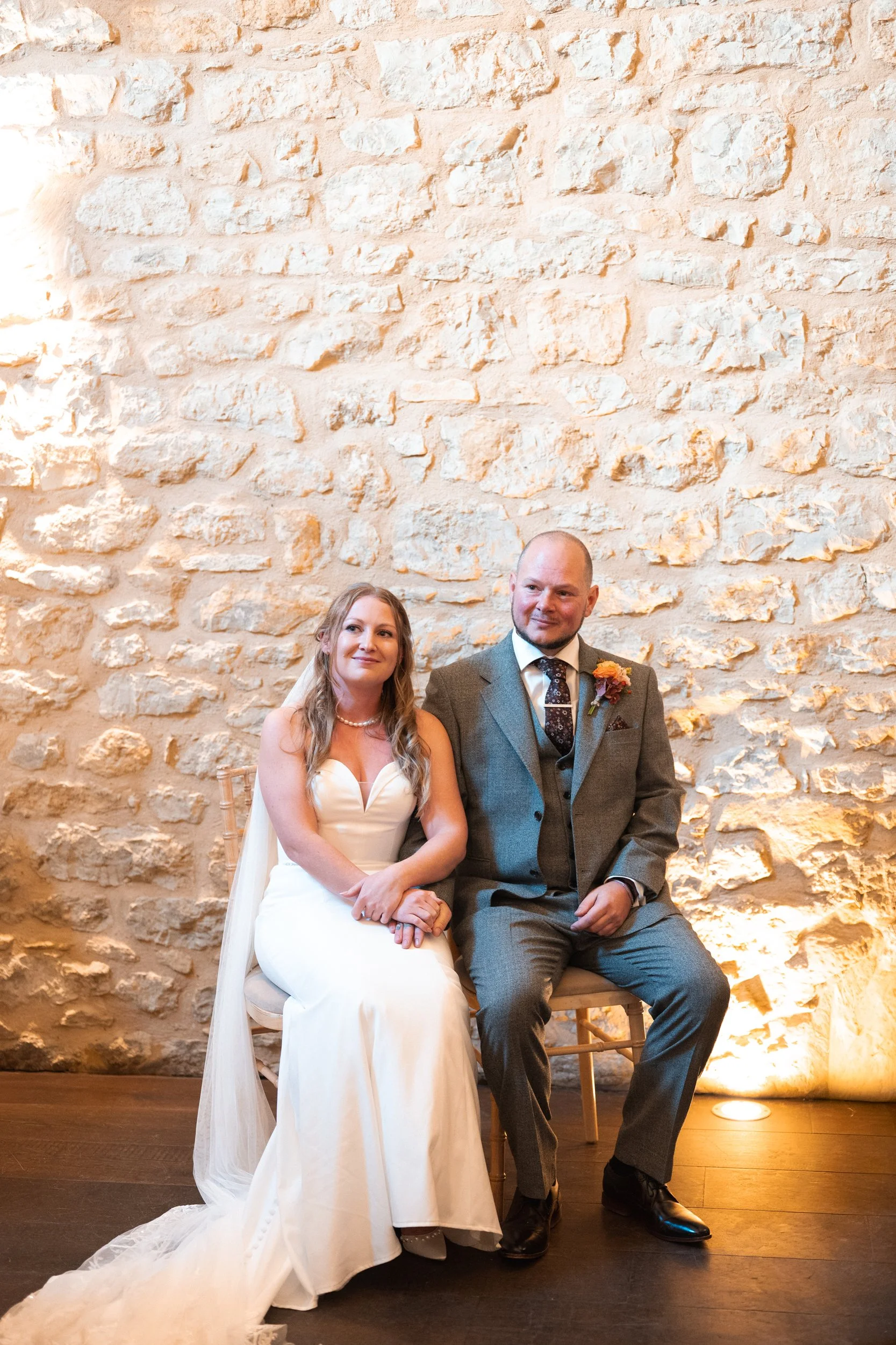 A bride and groom sitting together on a chair against a textured stone wall at their wedding reception.