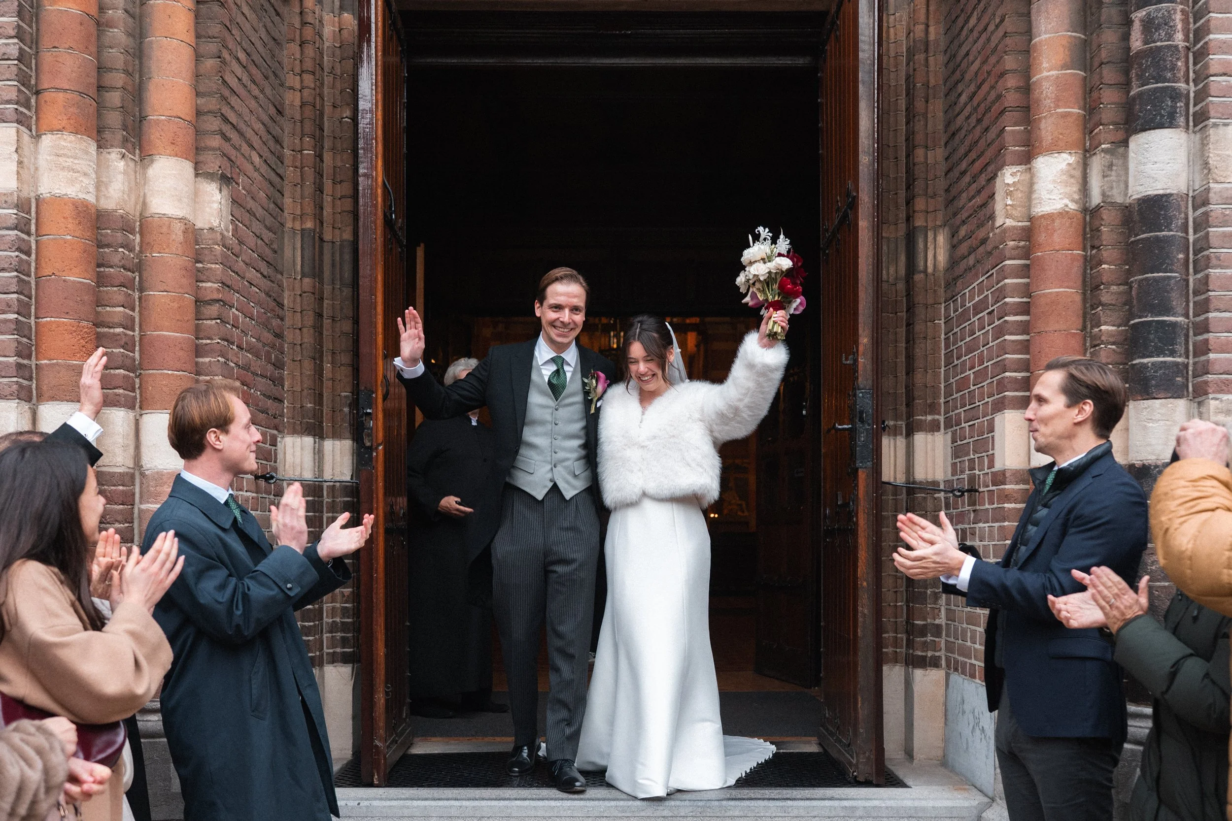 Bride and groom exit a church, smiling and raising their hands, as friends or family claps and celebrates outside the entrance.