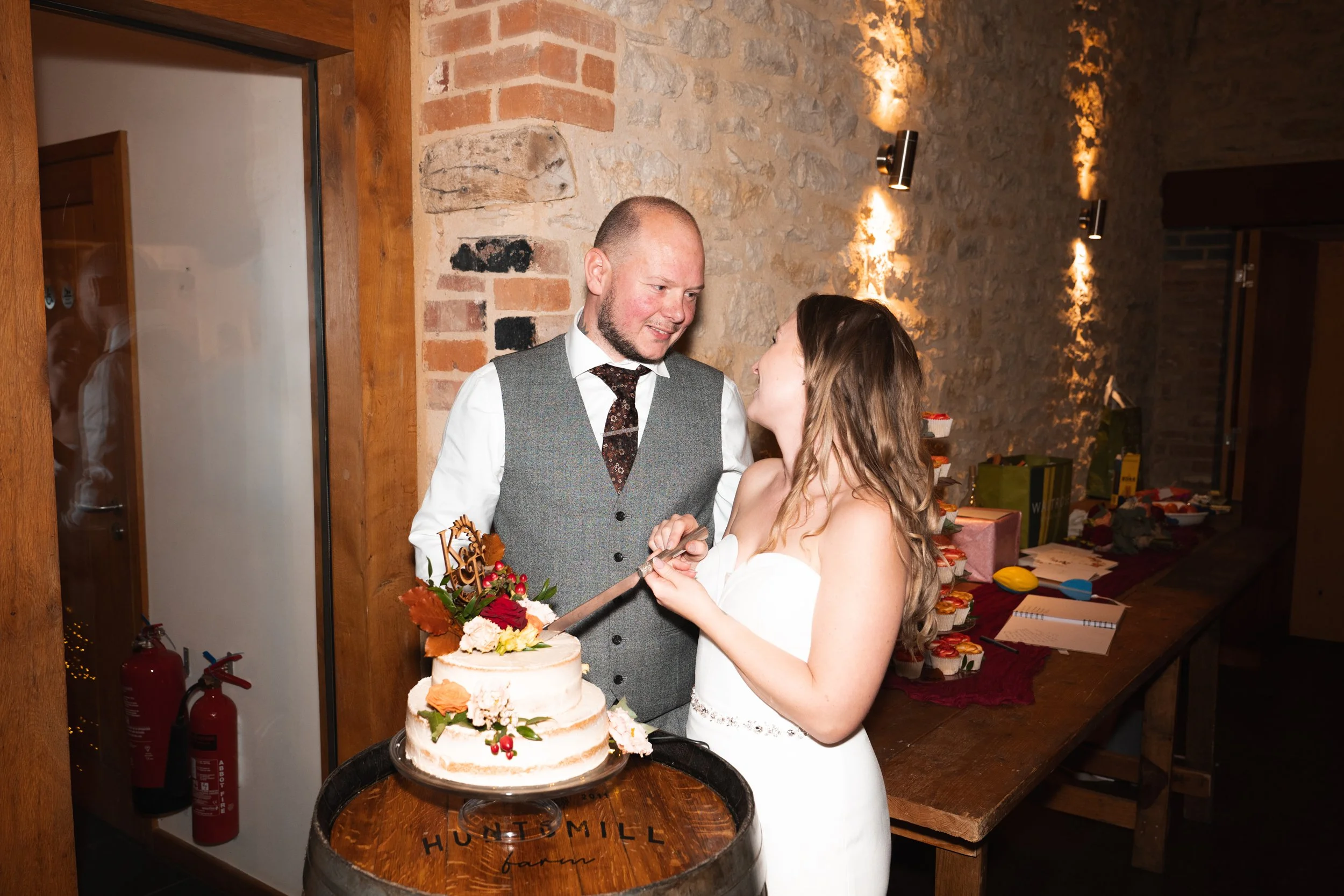 A bride and groom cutting their wedding cake together in a rustic reception hall with exposed brick and stone walls, surrounded by party favors and decorations.