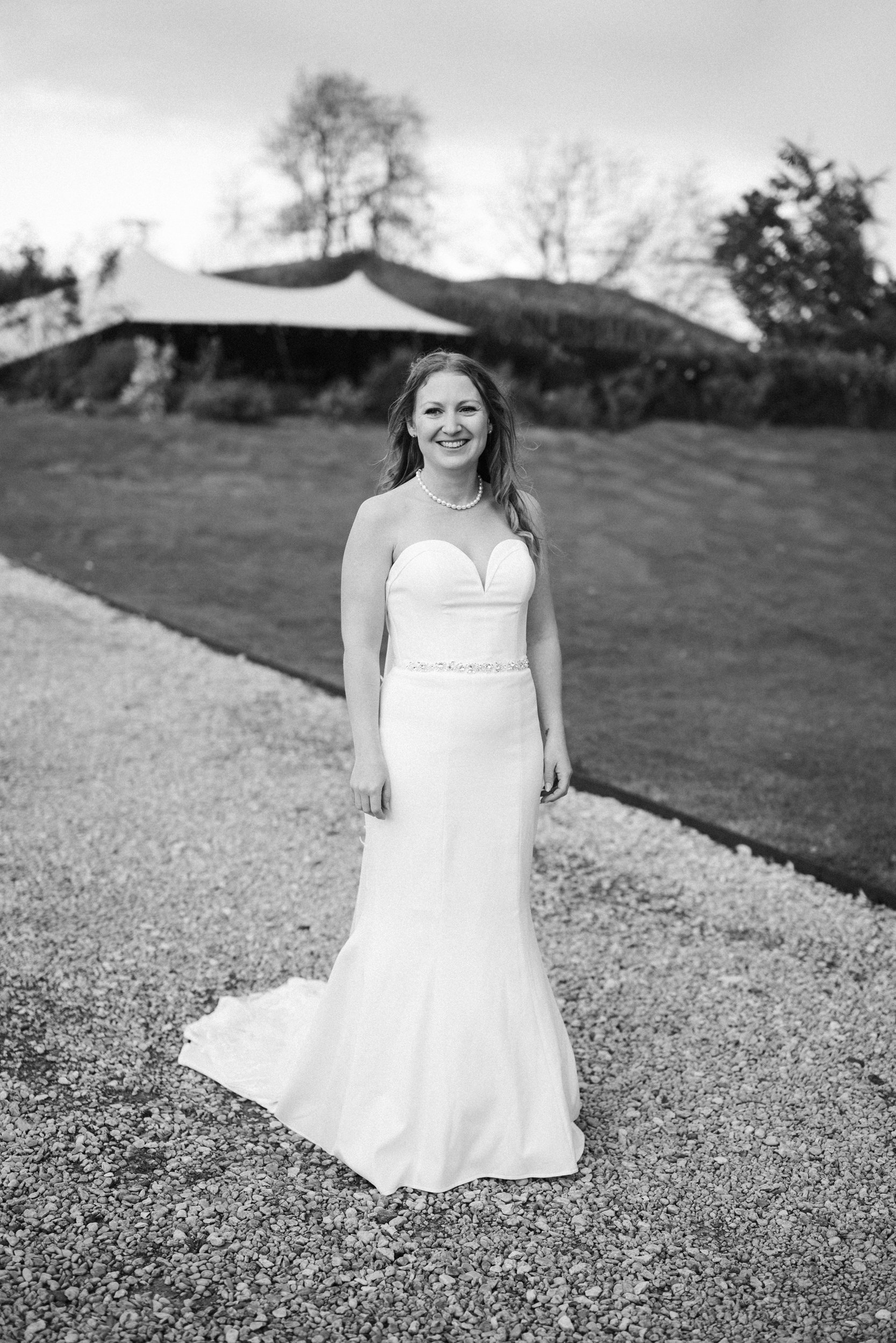 A smiling woman in a strapless wedding dress standing outdoors on a gravel path with a grassy hill and trees in the background.