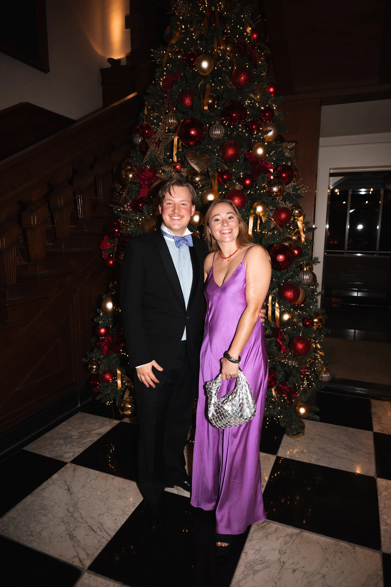A man in a black suit and a woman in a purple dress standing in front of a decorated Christmas tree with red and gold ornaments.