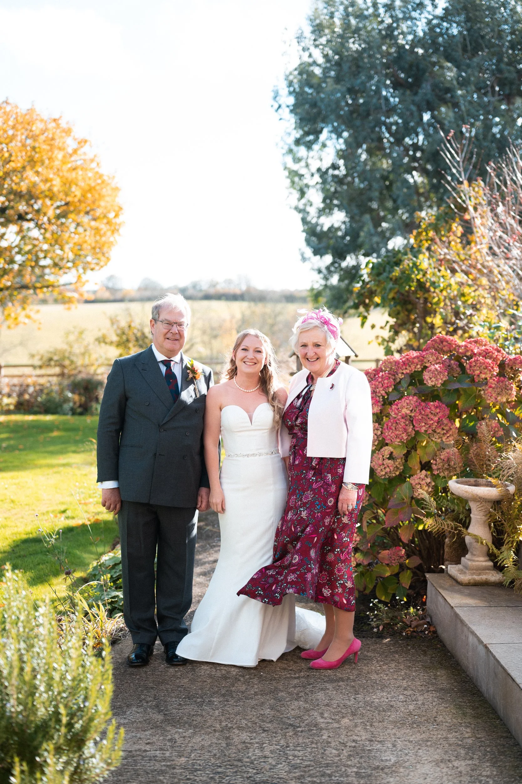 A bride in a white wedding gown standing between an older man in a dark suit and an older woman in a floral dress and pink shoes outdoors on a sunny day, with trees and bushes in the background.