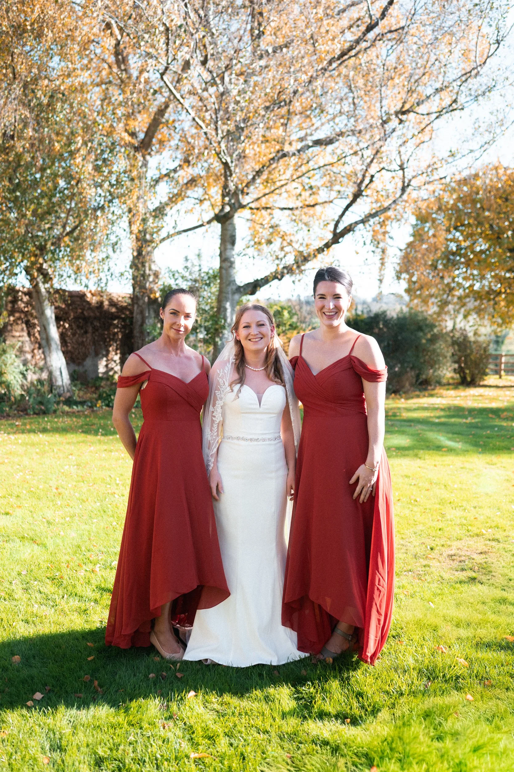 Three women, a bride in a white wedding dress with a veil and two bridesmaids in red dresses, standing outdoors on a sunny day with fall foliage in the background.