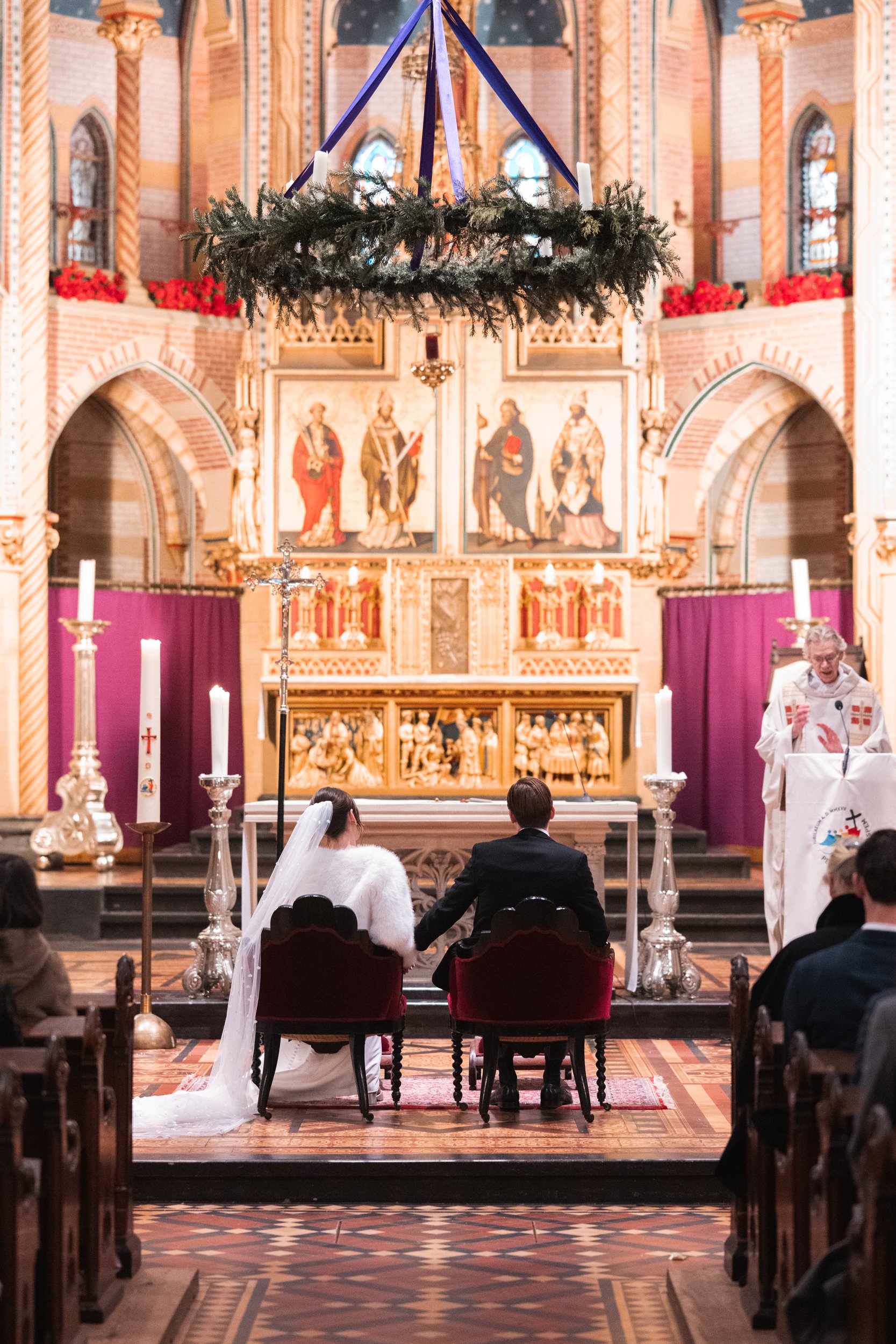 A wedding ceremony taking place inside a decorated church. The bride and groom are sitting together in front of the altar, holding hands. The bride is wearing a white dress and veil, and the groom is in a black suit. There are candles and religious s