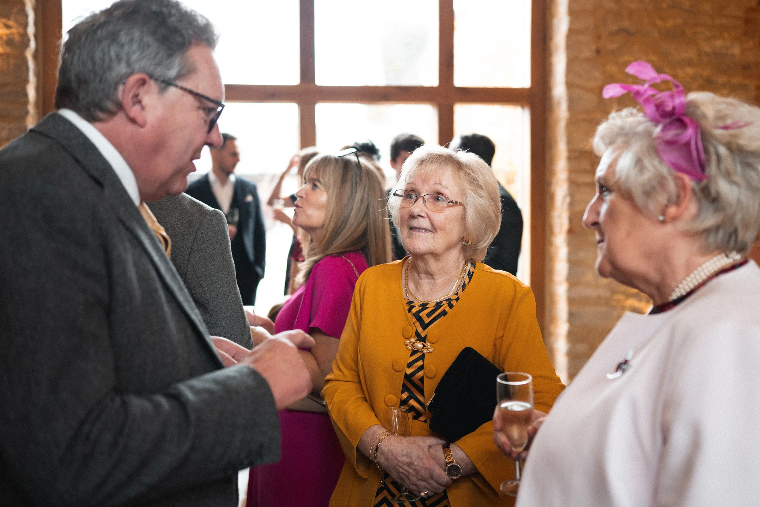 Group of elderly and middle-aged people at social event, talking and holding drinks in a warmly lit room with large window and brick wall background.