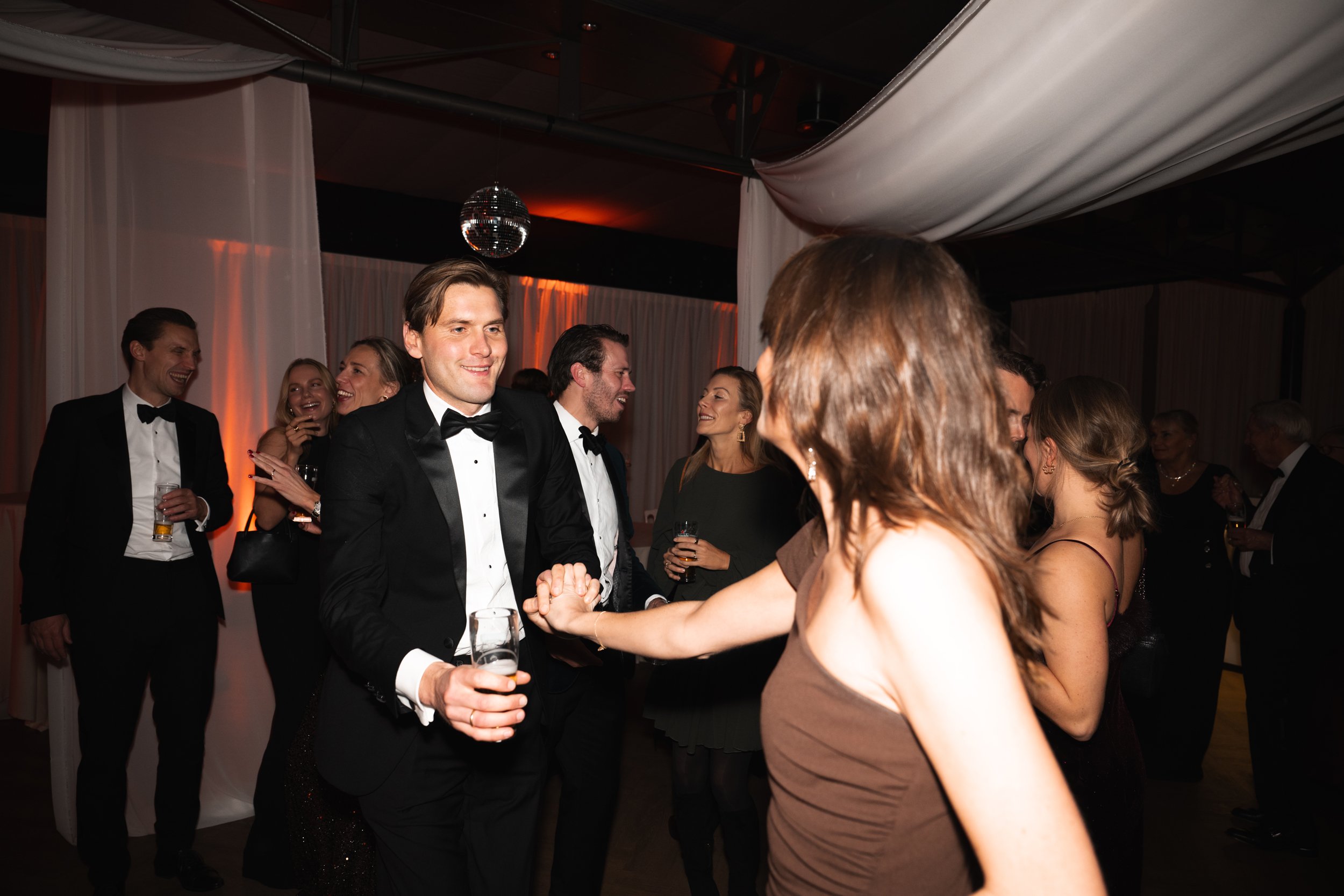 People in formal attire at a party, dancing and socializing indoors, with a disco ball overhead.