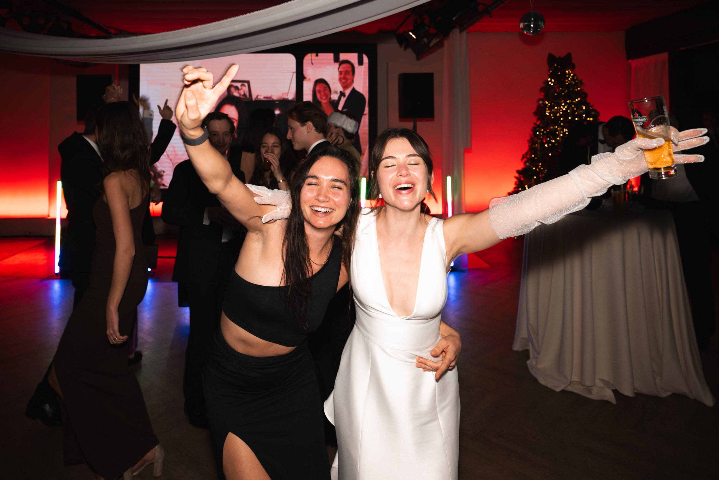 Two women celebrating at a party, smiling with arms raised, one holding a drink, at a decorated indoor event with a Christmas tree and other guests in the background.
