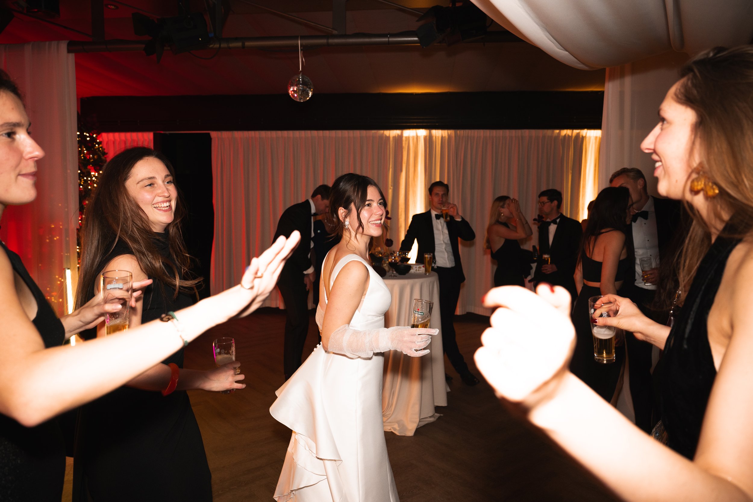 People in formal attire socializing at a wedding reception, with the bride in a white gown holding a glass and others holding drinks, in a decorated banquet hall.