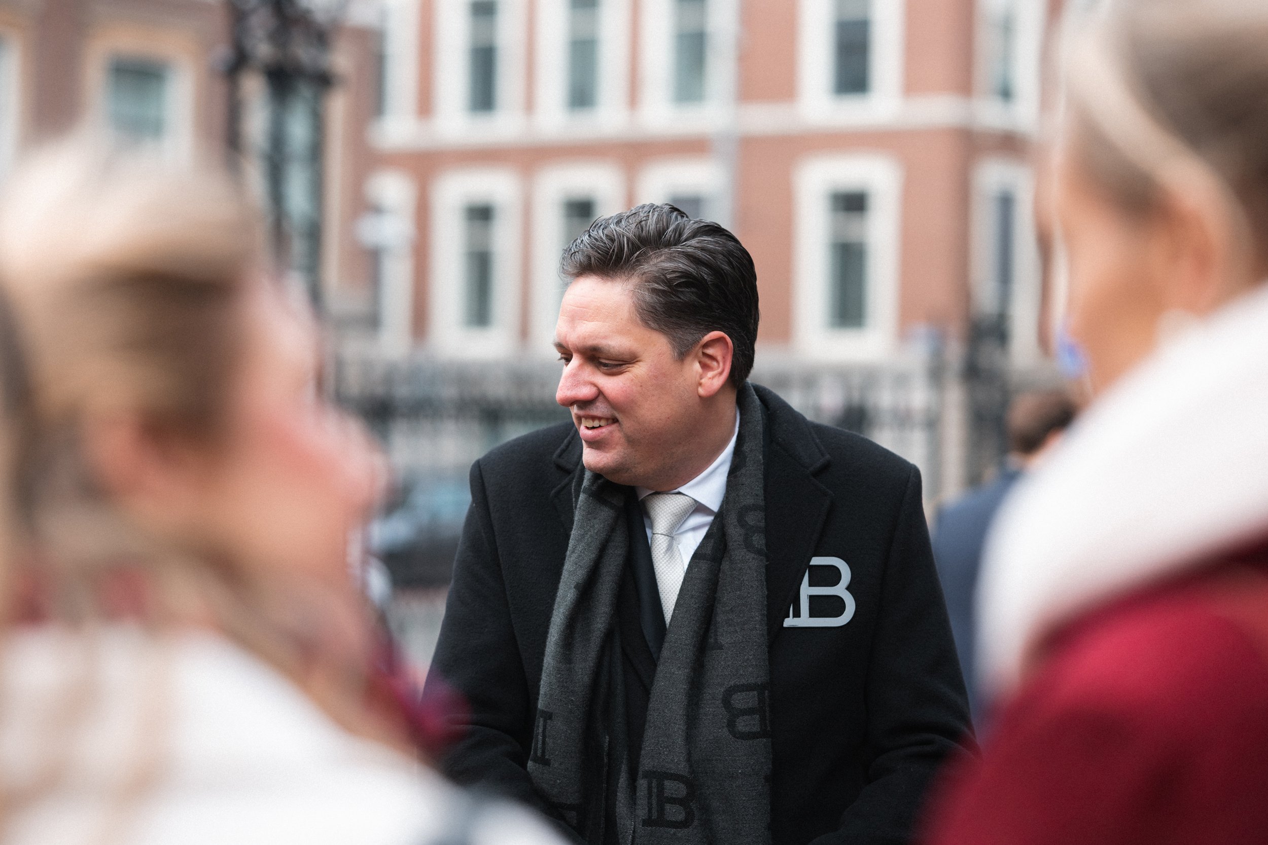 Man in a black coat and white tie talking to two women in coats outdoors, with a brick building visible in the background.