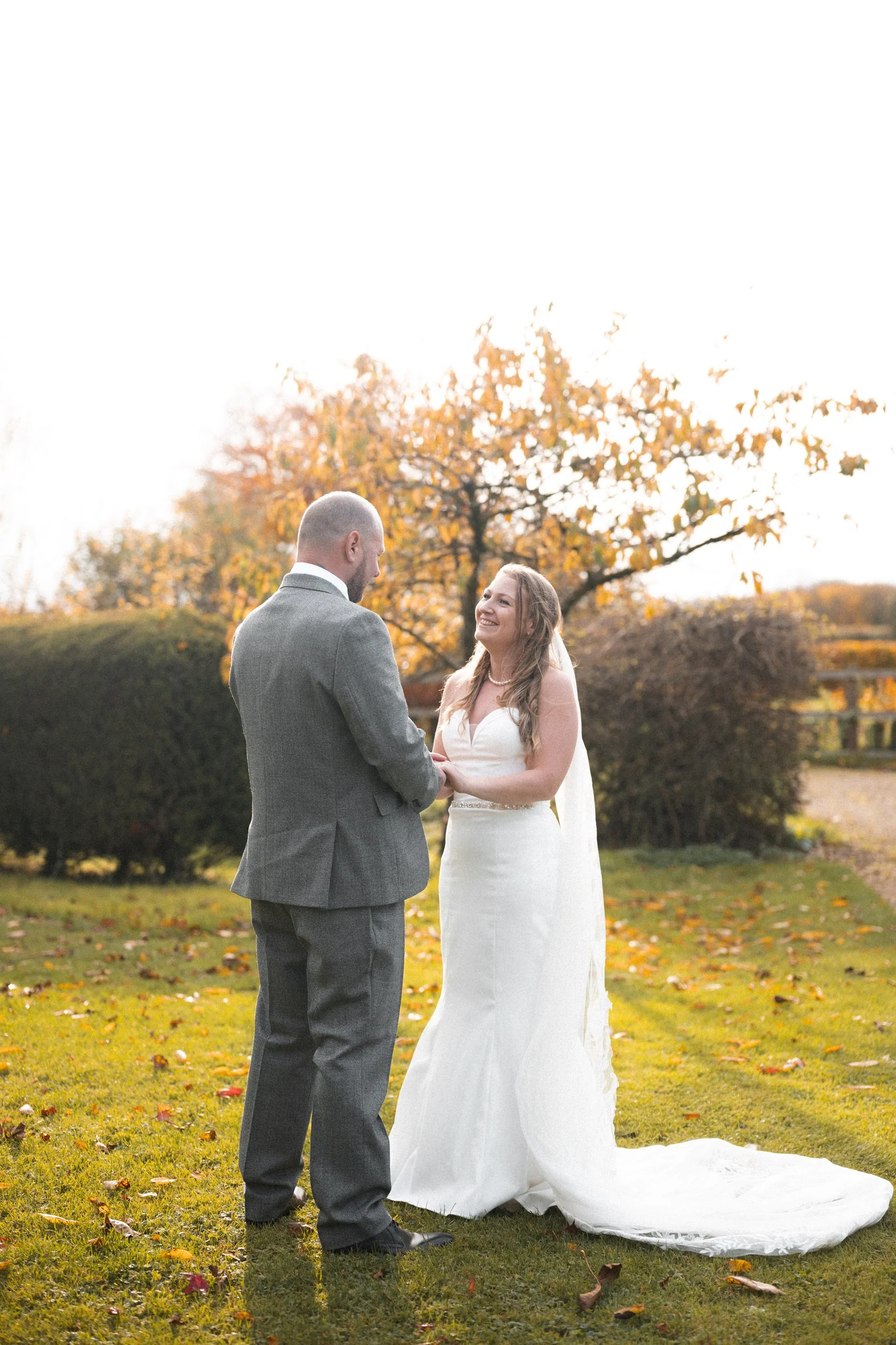 A bride and groom standing outdoors on a grassy area during autumn, holding hands and smiling at each other, with trees and a wooden fence in the background.