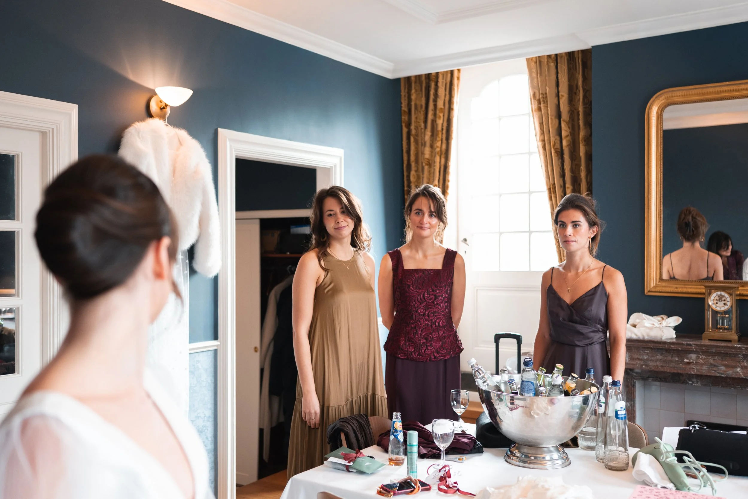 Three women in elegant dresses standing near a table with various drinks and items inside a decorated room, with a woman with her hair in an updo visible in the foreground, and a large mirror and window in the background.