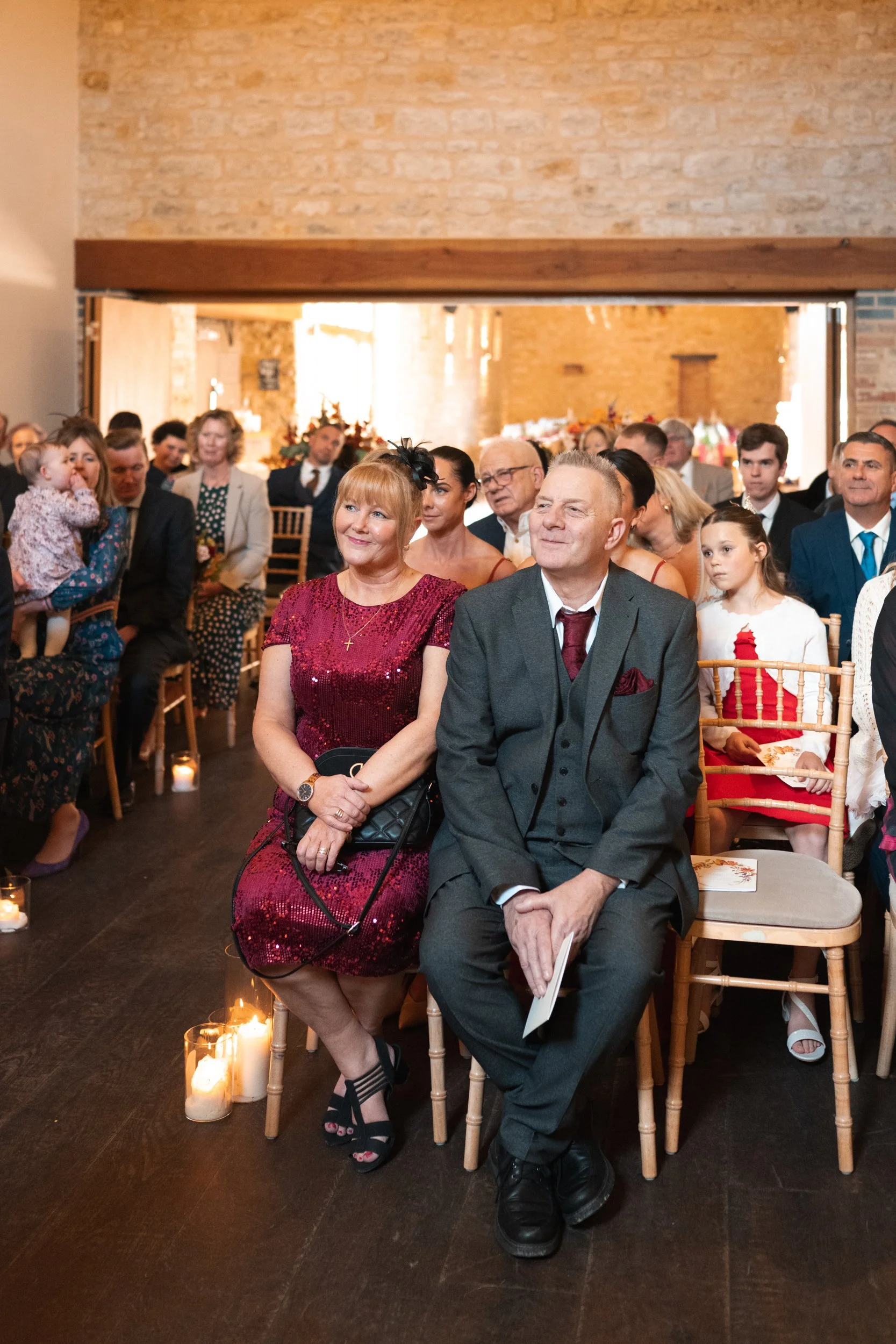 People attending a formal event seated in a rustic venue with exposed brick walls. Some are dressed in elegant attire, including a woman in a red sequin dress and a man in a gray suit, sitting in the front row. Candles line the floor near their seats