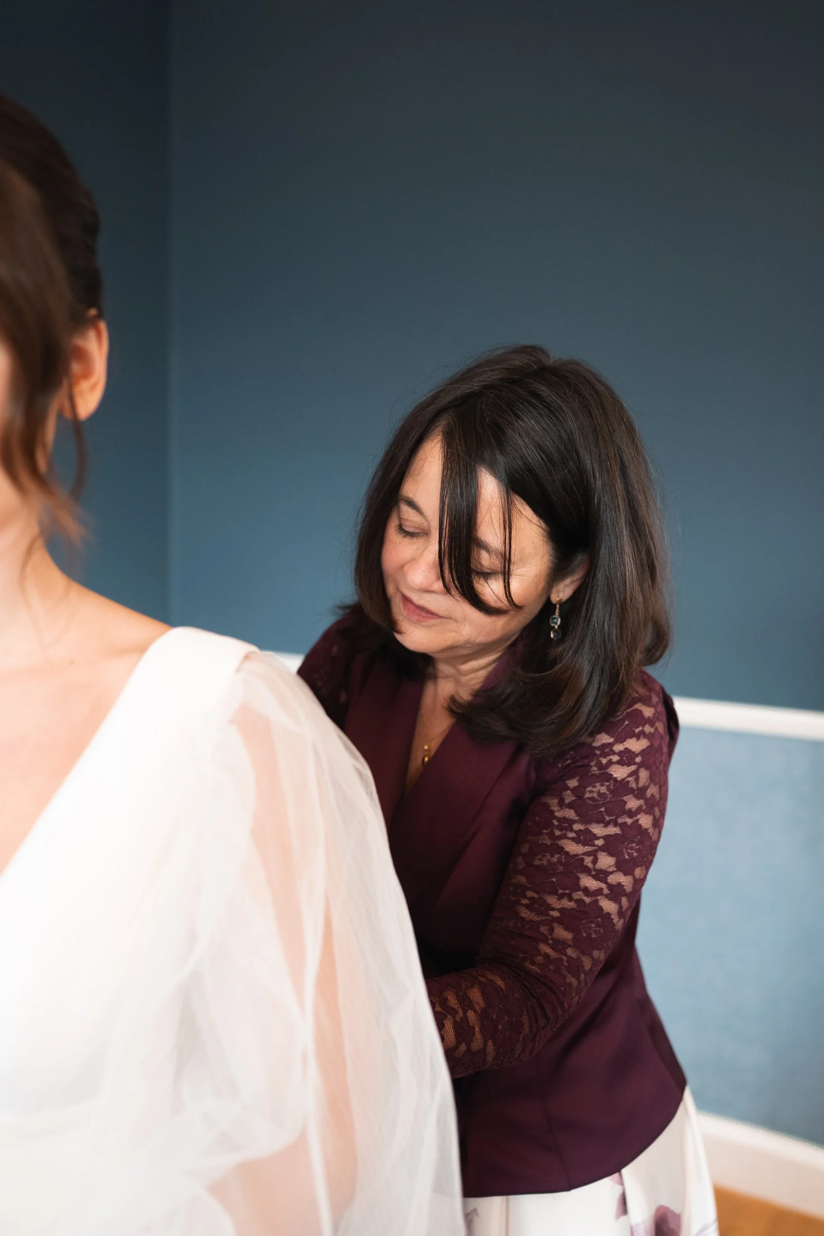A woman with dark hair, wearing a burgundy lace top, helping another woman in a white dress, with a blue wall in the background.