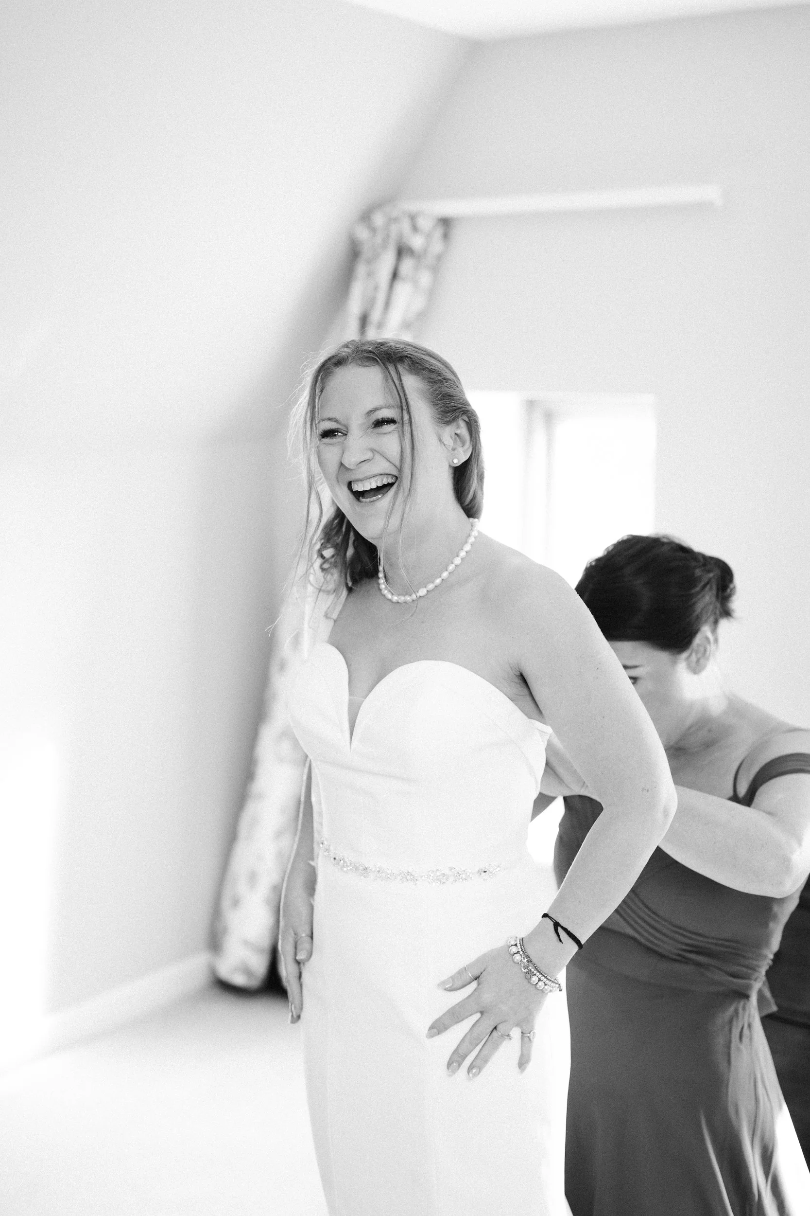 A joyful bride in a strapless white wedding dress being helped by another woman in a sleeveless dress, with a window and floral curtains in the background.
